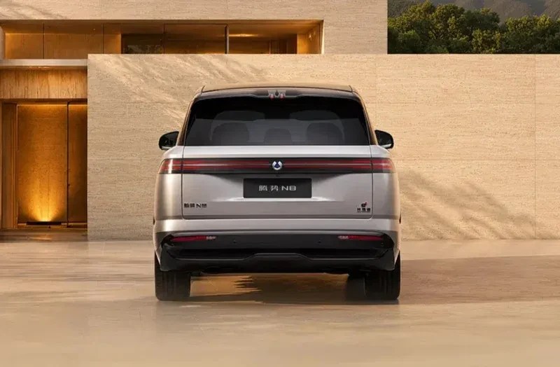 Traseira de um carro SUV cinza-prateado, com lanternas horizontais e um logotipo circular no centro, estacionado em frente a uma parede de pedra clara com uma entrada iluminada à esquerda e vegetação no topo à direita