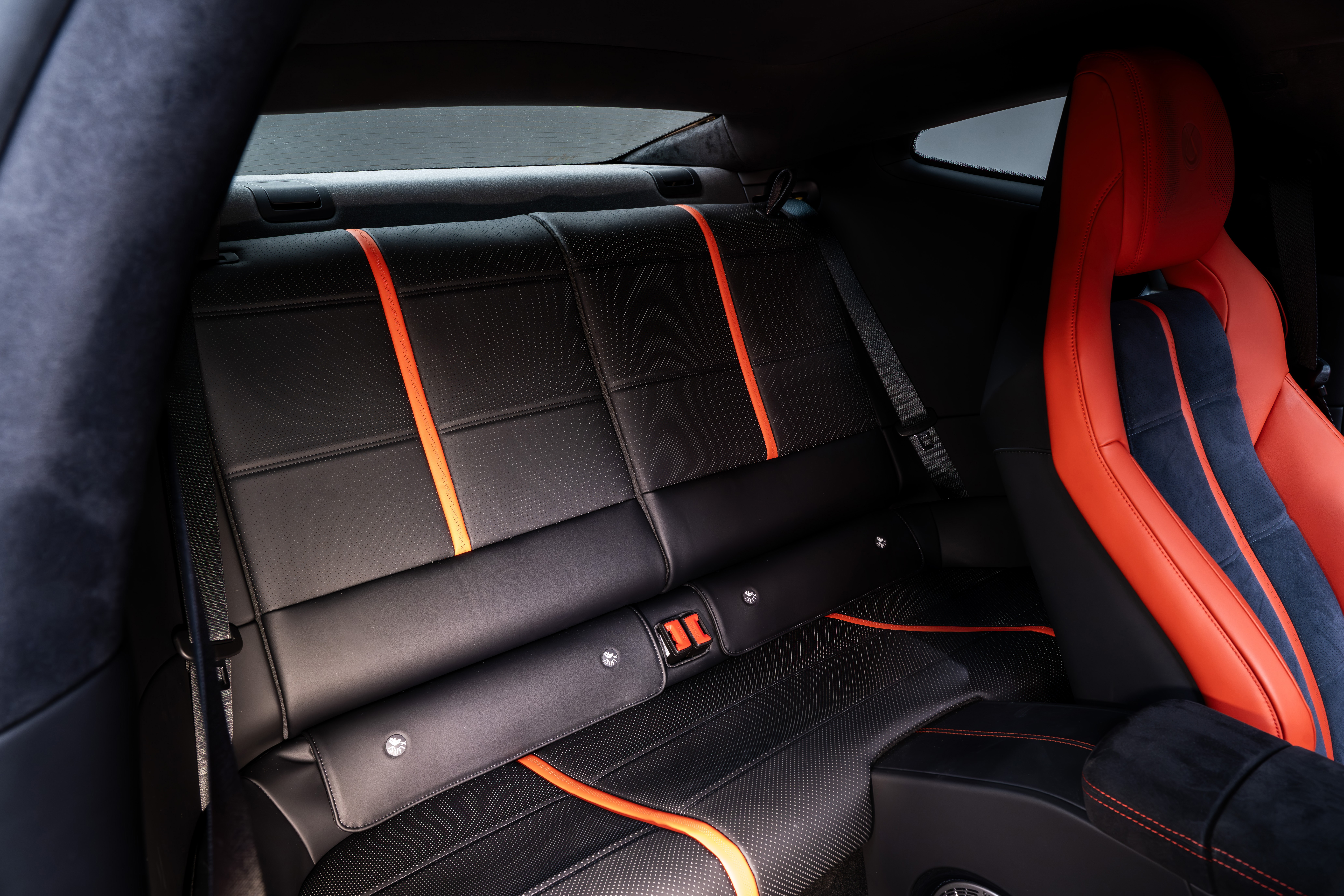 Interior de um carro esportivo com bancos traseiros pretos de couro, detalhados com faixas verticais e horizontais laranja. O banco dianteiro direito, visível parcialmente, é vermelho com detalhes em azul escuro. O cinto de segurança traseiro está visível, e o ambiente é escuro, com luz vindo das janelas