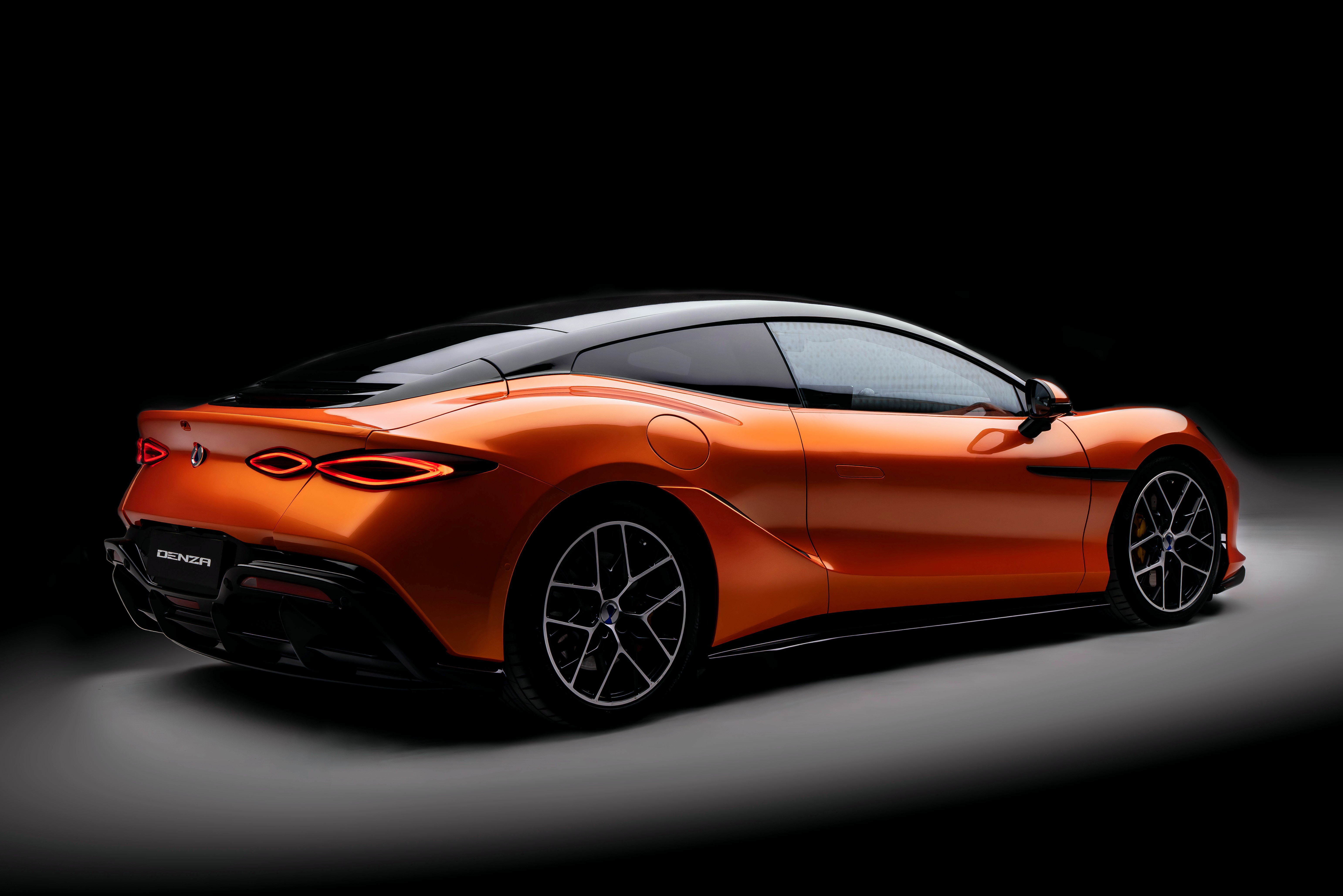 Carro esportivo laranja com teto preto, visto de tr&aacute;s para a direita, em um fundo escuro. As lanternas traseiras s&atilde;o finas e ovais, e as rodas t&ecirc;m design geom&eacute;trico