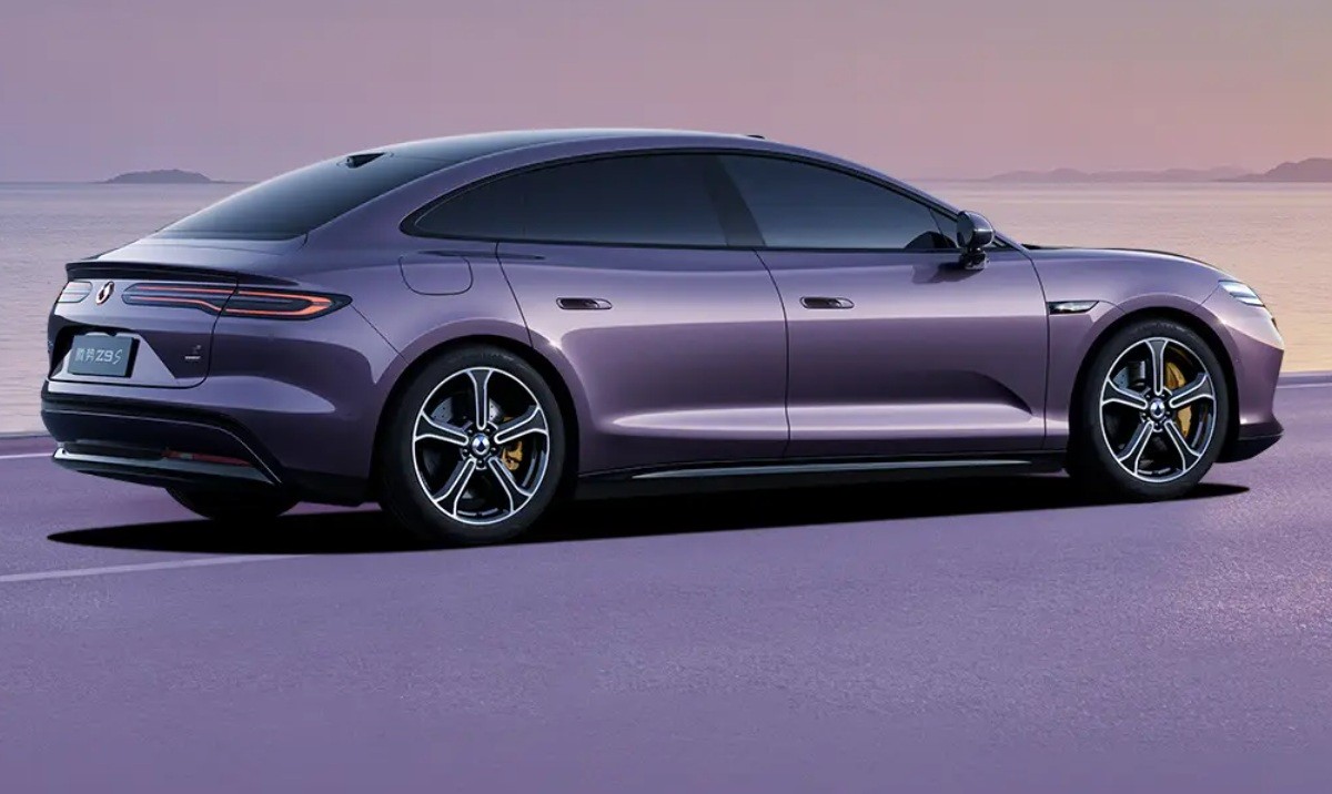 Carro elétrico roxo escuro, modelo sedan, estacionado em uma estrada roxa com o mar e montanhas ao fundo, sob um céu alaranjado ao pôr do sol
