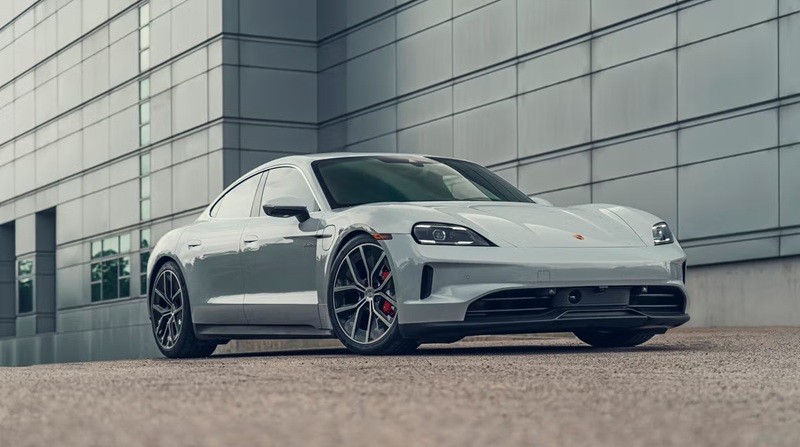 Carro esportivo Porsche Taycan cinza claro, elétrico, estacionado em frente a um edifício moderno com fachada de painéis metálicos e janelas retangulares, em um dia claro