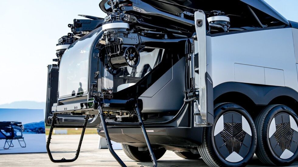 Veículo futurista preto e prata, com cabine de vidro e hélices visíveis na parte superior, em um ambiente externo com céu claro. O veículo possui rodas com design inovador, e uma estrutura de pouso preta se estende da cabine. Ao fundo, uma imagem de montanhas e um lago.