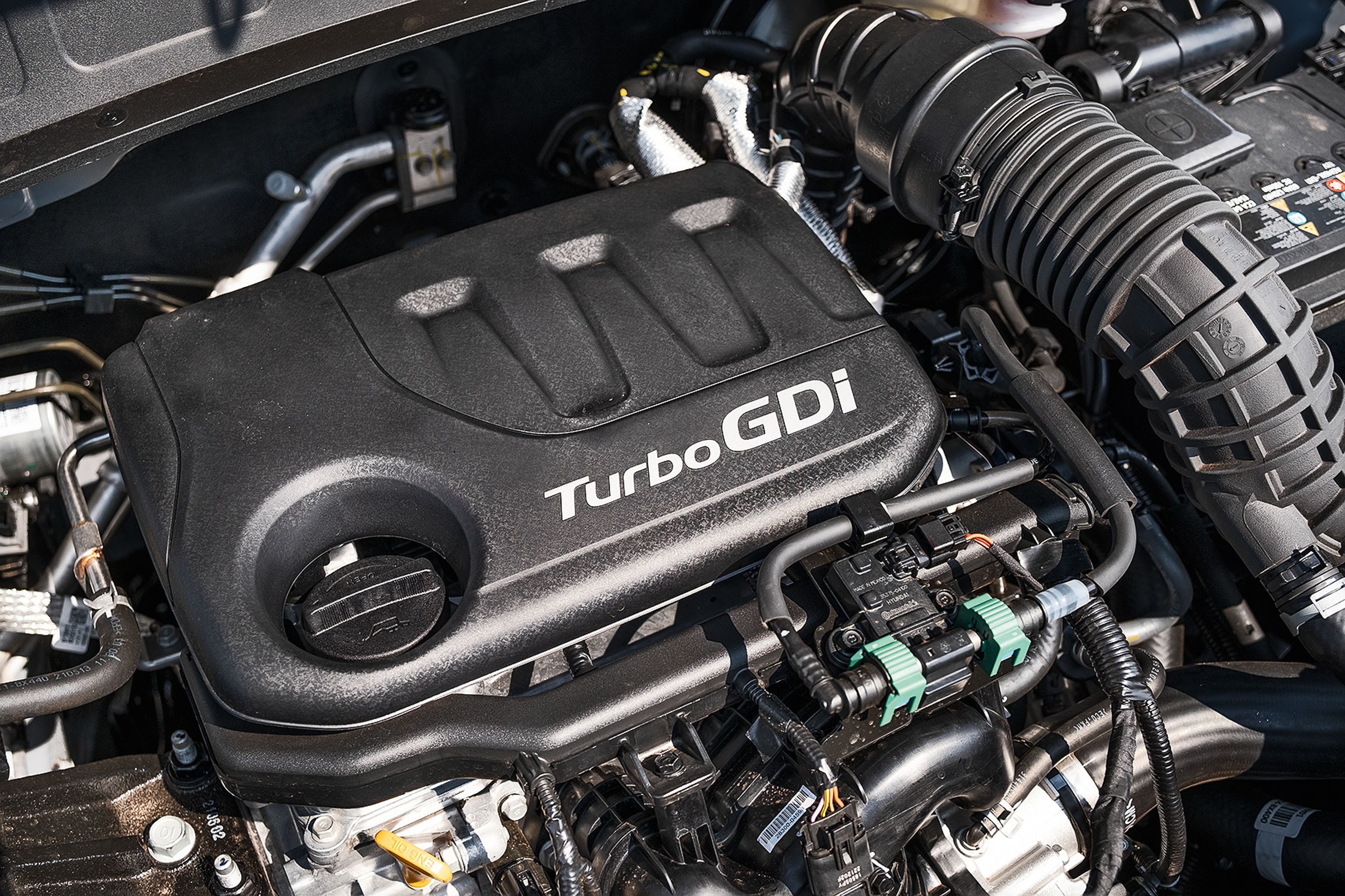 Motor de carro com a tampa preta do motor em destaque, exibindo o texto TurboGDi em branco. Ao redor, tubulações, fios e componentes mecânicos prateados e pretos, com alguns detalhes em verde e amarelo, sob luz forte