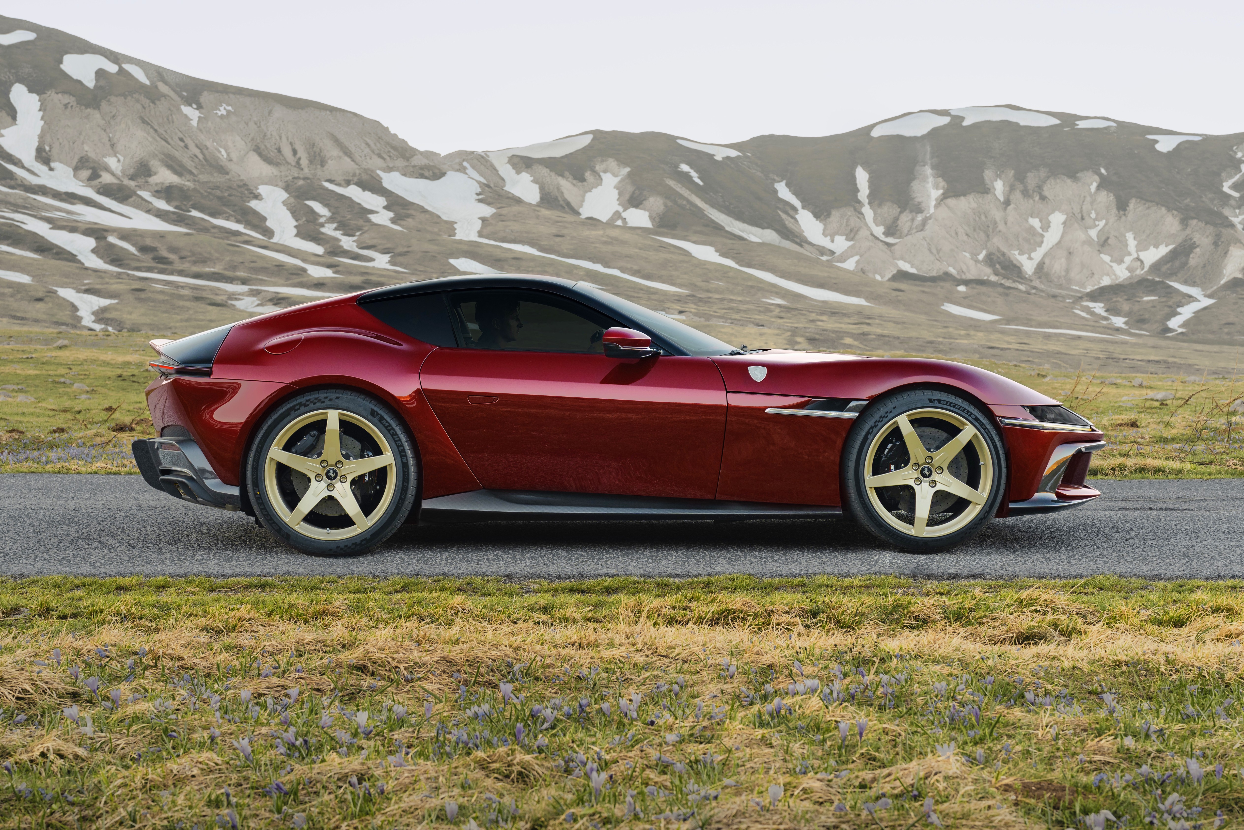 Carro esportivo vermelho escuro com teto preto e rodas douradas de cinco raios, estacionado em uma estrada asfaltada. Ao fundo, montanhas com picos nevados e vegetação rasteira com flores roxas no primeiro plano