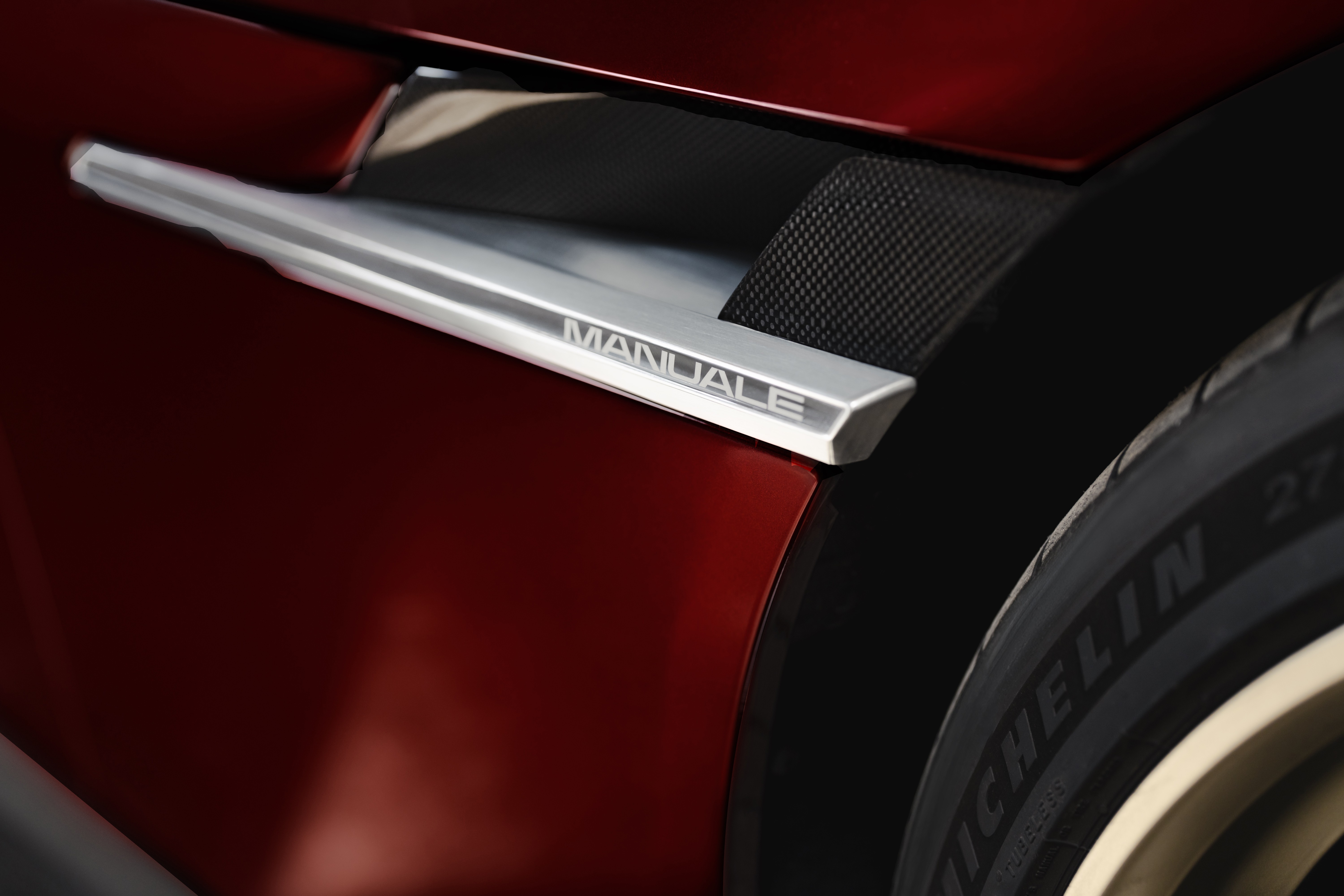 Detalhe da lateral de um carro esportivo vermelho-escuro, com um emblema prateado escrito MANUALE e uma grade preta texturizada. Parte de um pneu Michelin é visível à direita
