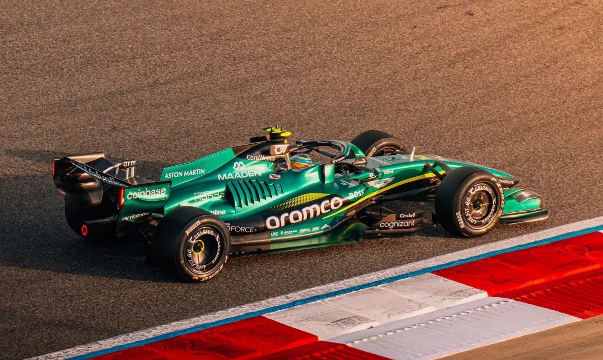 Carro de Fórmula 1 Aston Martin verde-esmeralda com detalhes em amarelo e preto, patrocinadores Aramco e Cognizant, em uma pista de corrida com zebras vermelhas e brancas, sob luz solar