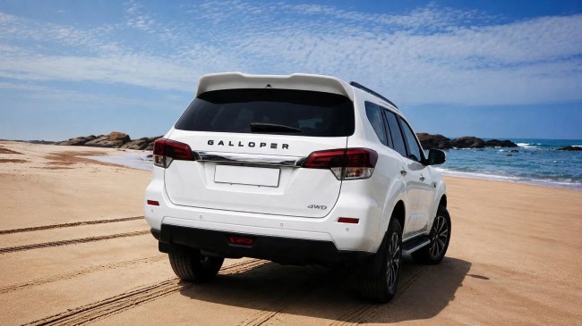 SUV branco Galloper estacionado na areia da praia, com o mar azul e rochas ao fundo sob um céu claro