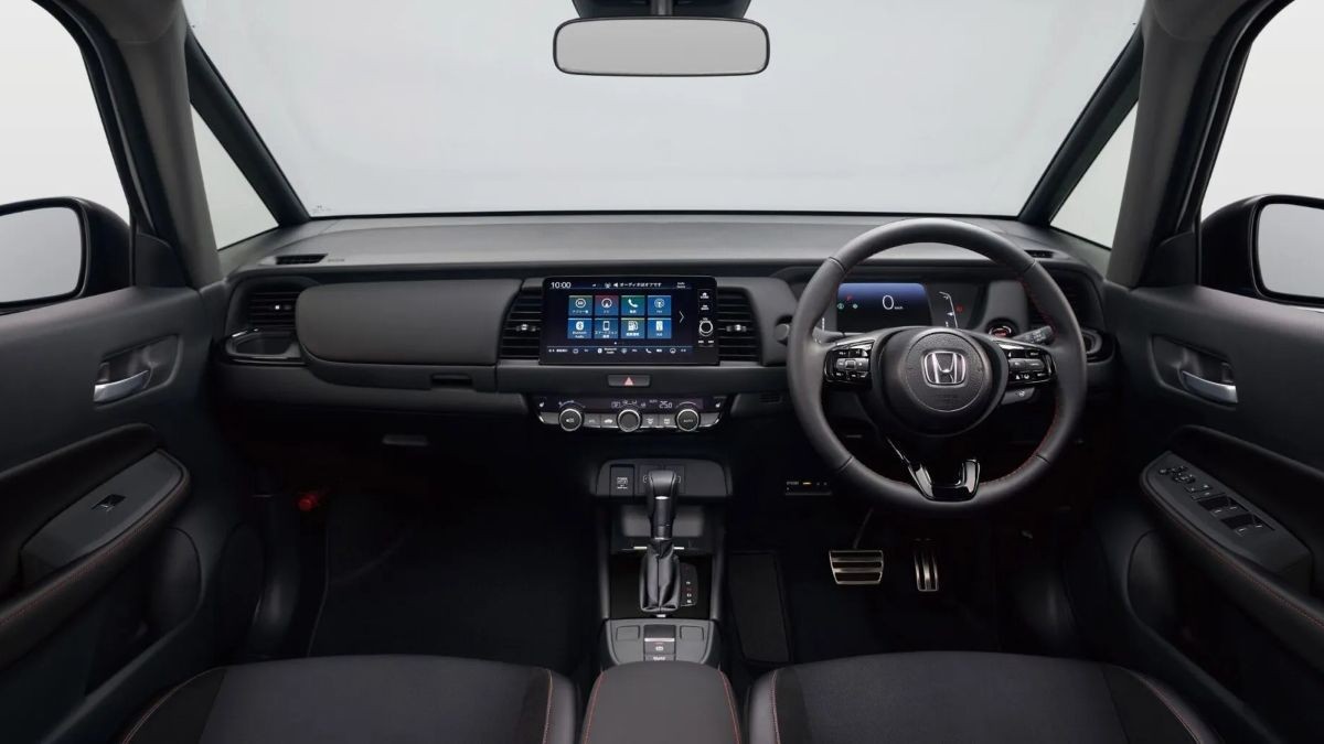 Interior de um carro moderno, com painel escuro e tela multimídia central exibindo ícones de aplicativos. Volante multifuncional com o logo da Honda, câmbio automático e pedais metálicos. Bancos dianteiros pretos com detalhes em vermelho, visíveis em primeiro plano