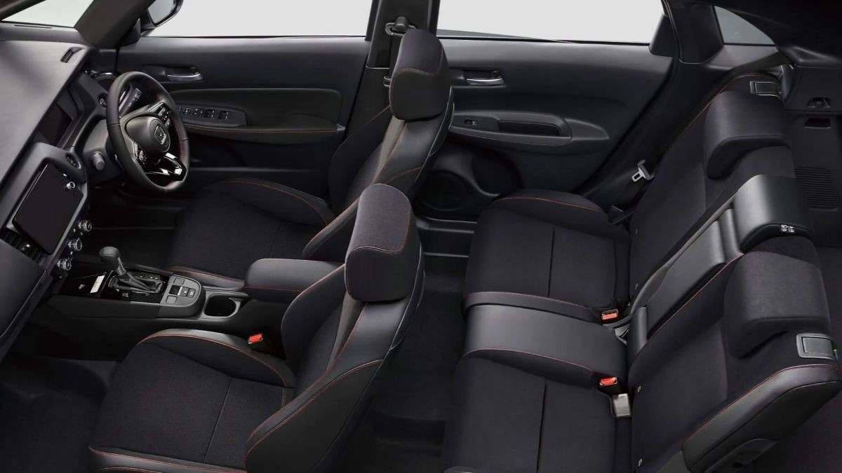 Interior de carro com bancos pretos e costura vermelha, volante, painel digital e câmbio automático visíveis no lado esquerdo. Os bancos traseiros estão rebatidos, mostrando a versatilidade do espaço.
