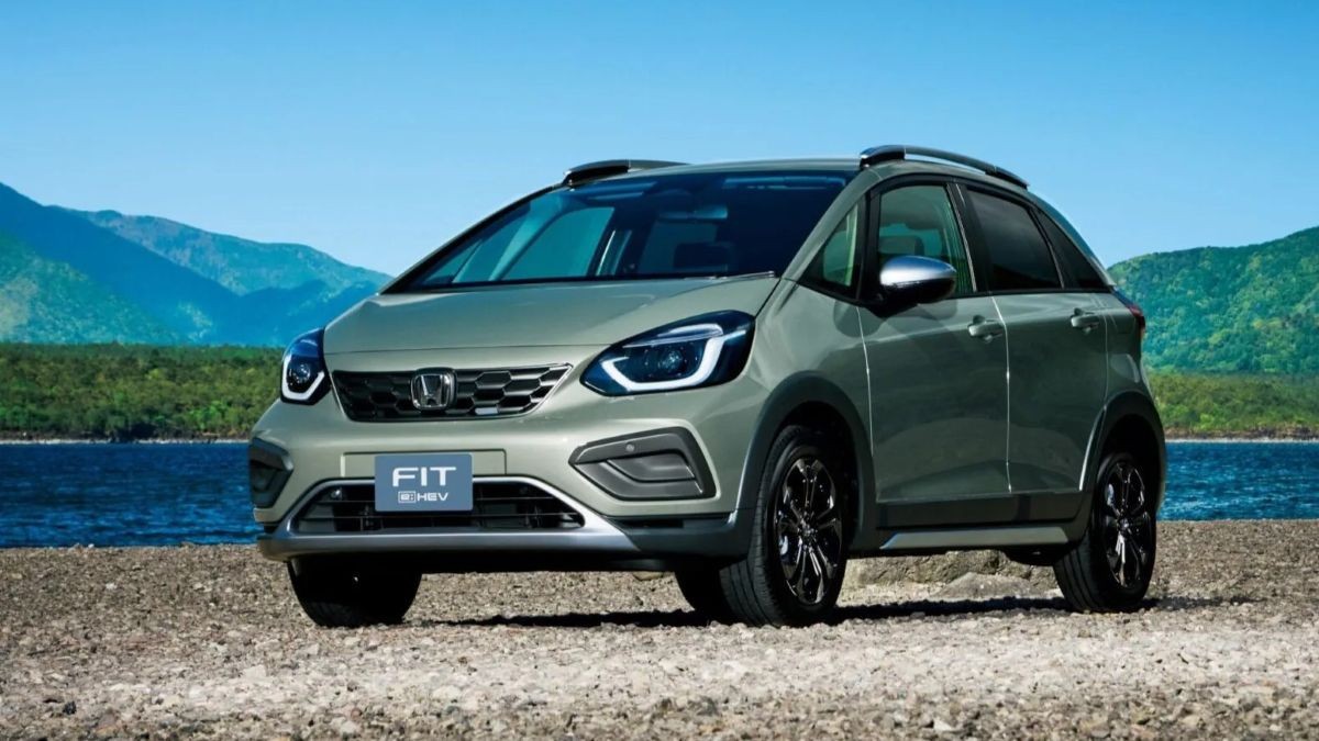 Carro Honda Fit e:HEV verde-oliva com detalhes pretos e rack de teto, estacionado em terreno de cascalho, com montanhas verdes e um lago azul ao fundo sob céu claro