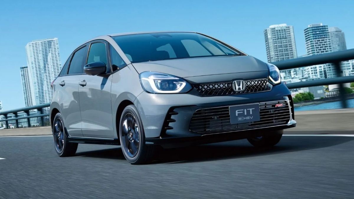 Carro Honda Fit cinza-claro em movimento, visto de frente e levemente de lado, com prédios altos e um rio ao fundo sob céu azul