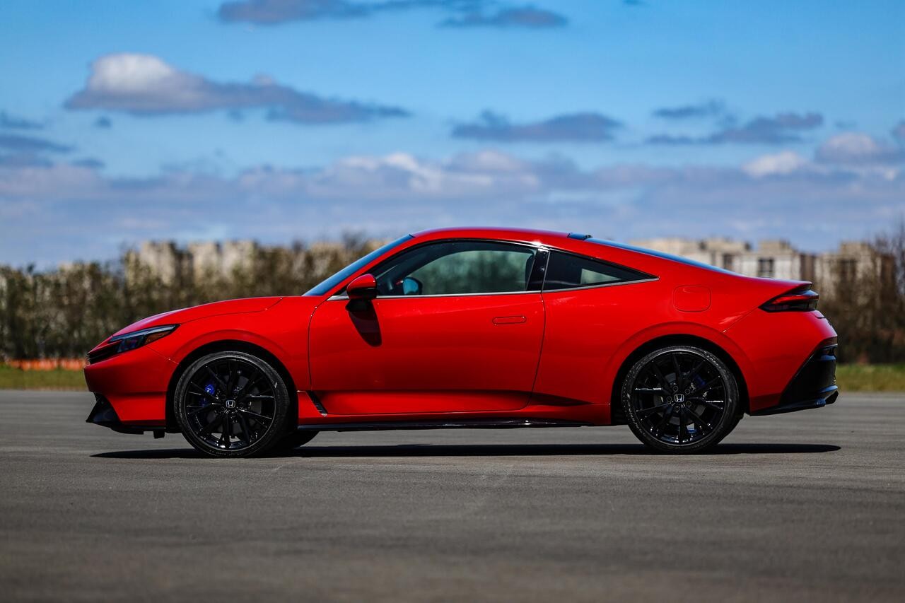 Carro esportivo vermelho brilhante, com teto solar e rodas pretas, estacionado em uma pista de asfalto sob céu azul com nuvens esparsas