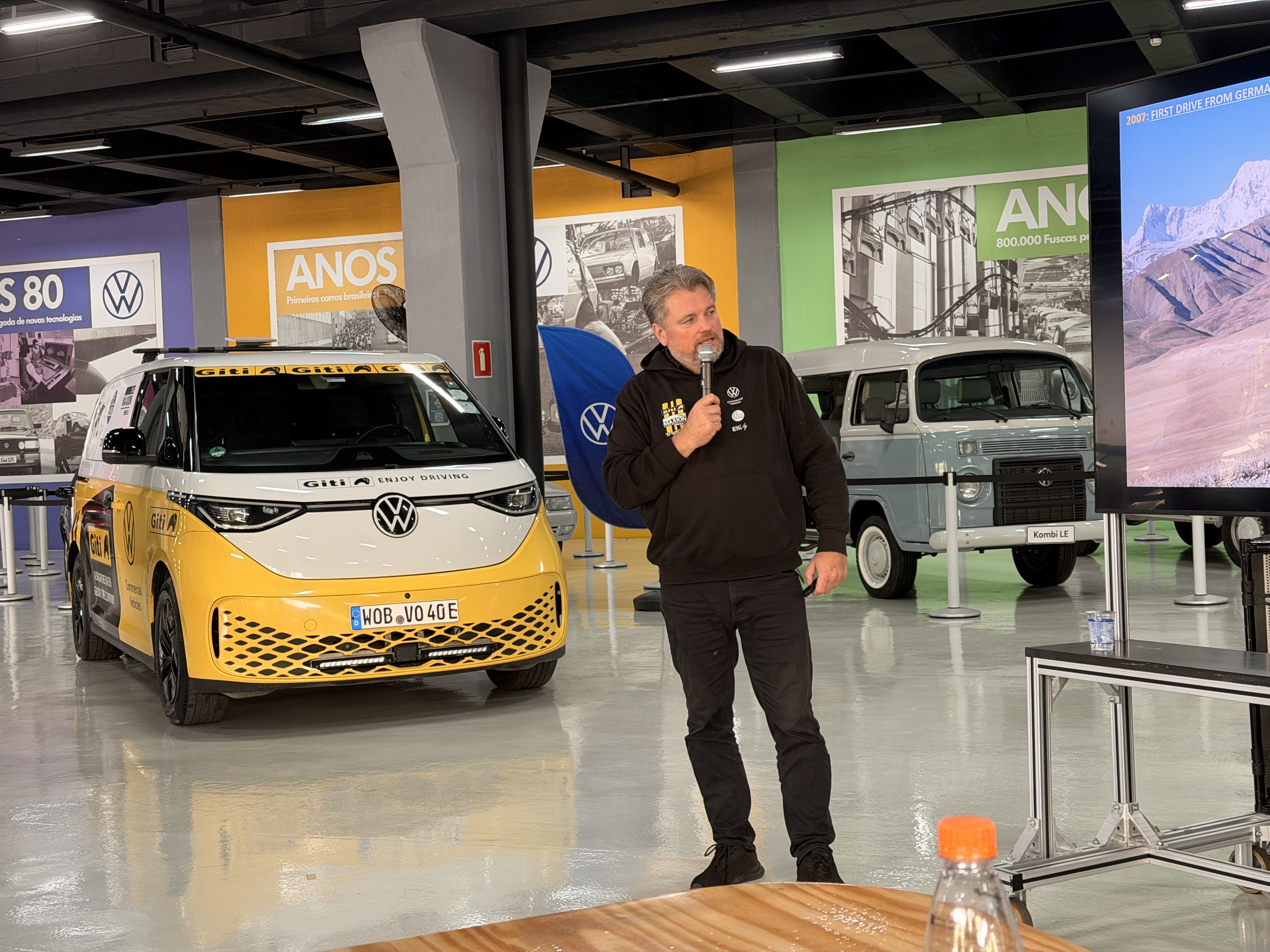 Homem de barba grisalha, vestindo moletom preto e calça escura, fala ao microfone em um evento. Atrás dele, uma van elétrica branca e amarela, e uma Kombi azul-clara antiga. Ao fundo, painéis com fotos de carros e um telão exibindo uma paisagem montanhosa