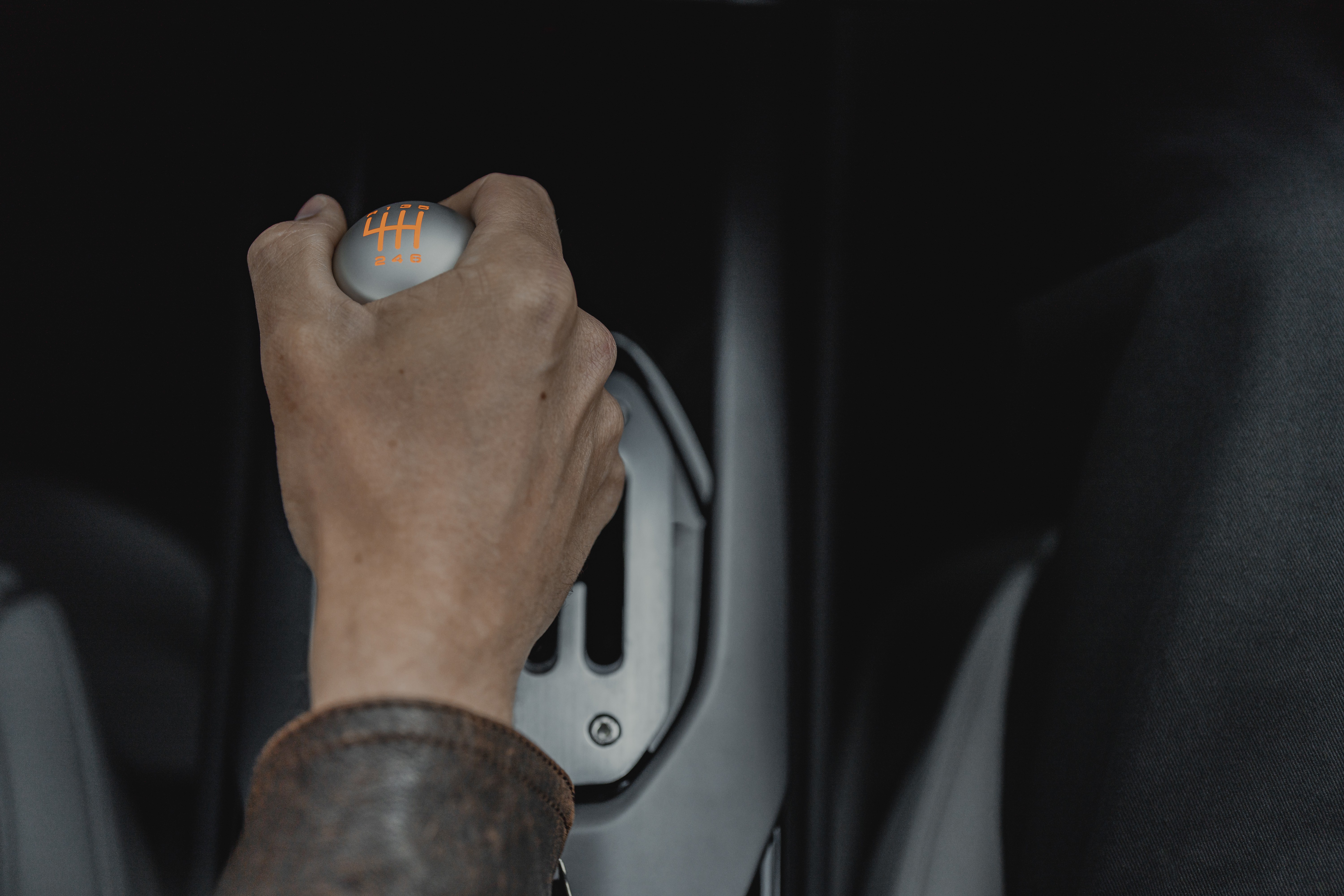 Mão de uma pessoa com jaqueta de couro marrom segurando o câmbio manual de um carro, com a manopla cinza e números de marcha laranja, em um interior escuro
