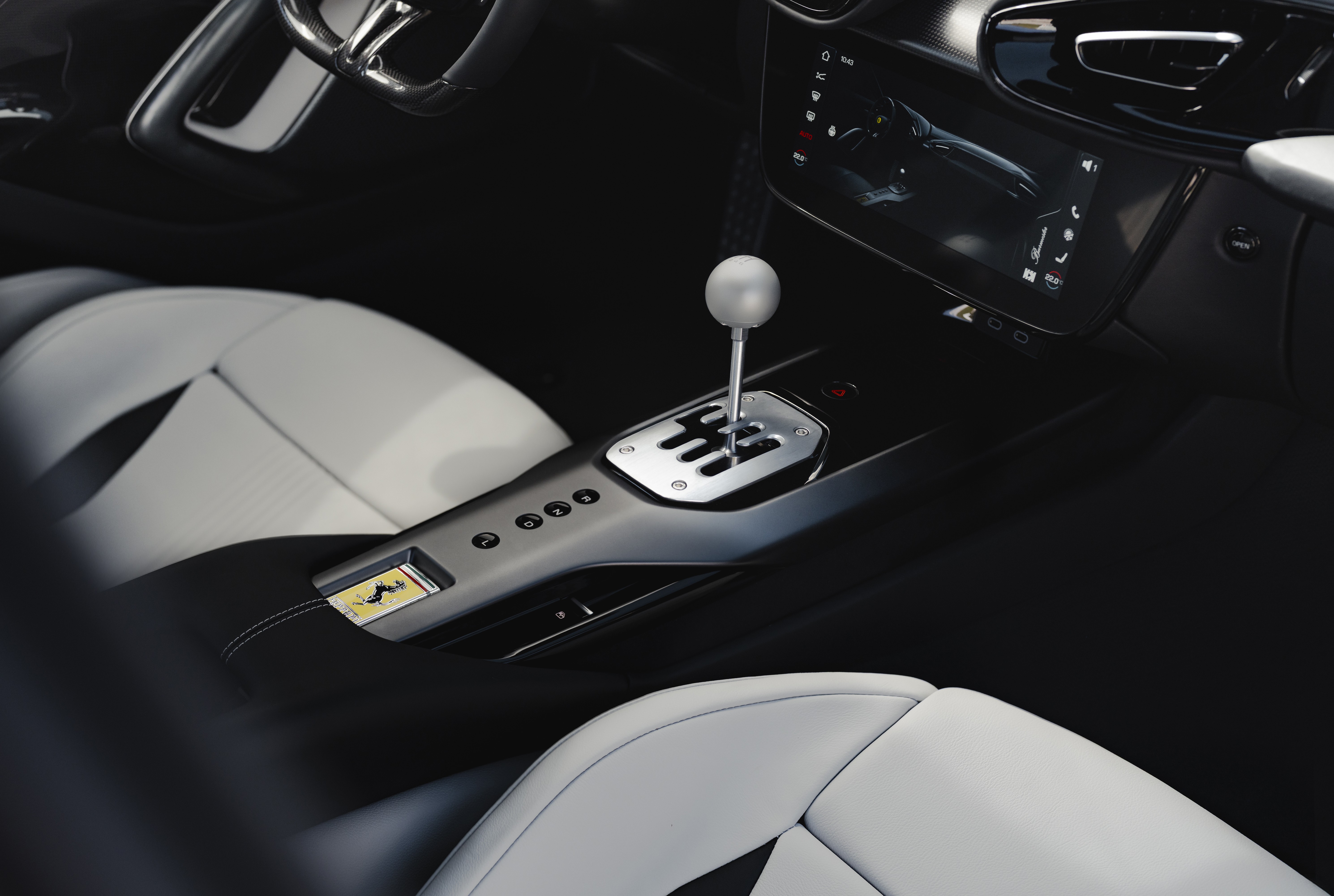 Interior de um carro esportivo com bancos de couro branco e preto, console central cinza escuro e câmbio manual cromado com manopla esférica. O painel possui uma tela multimídia e o volante é visível no canto superior esquerdo. No console, há um emblema dourado da Ferrari