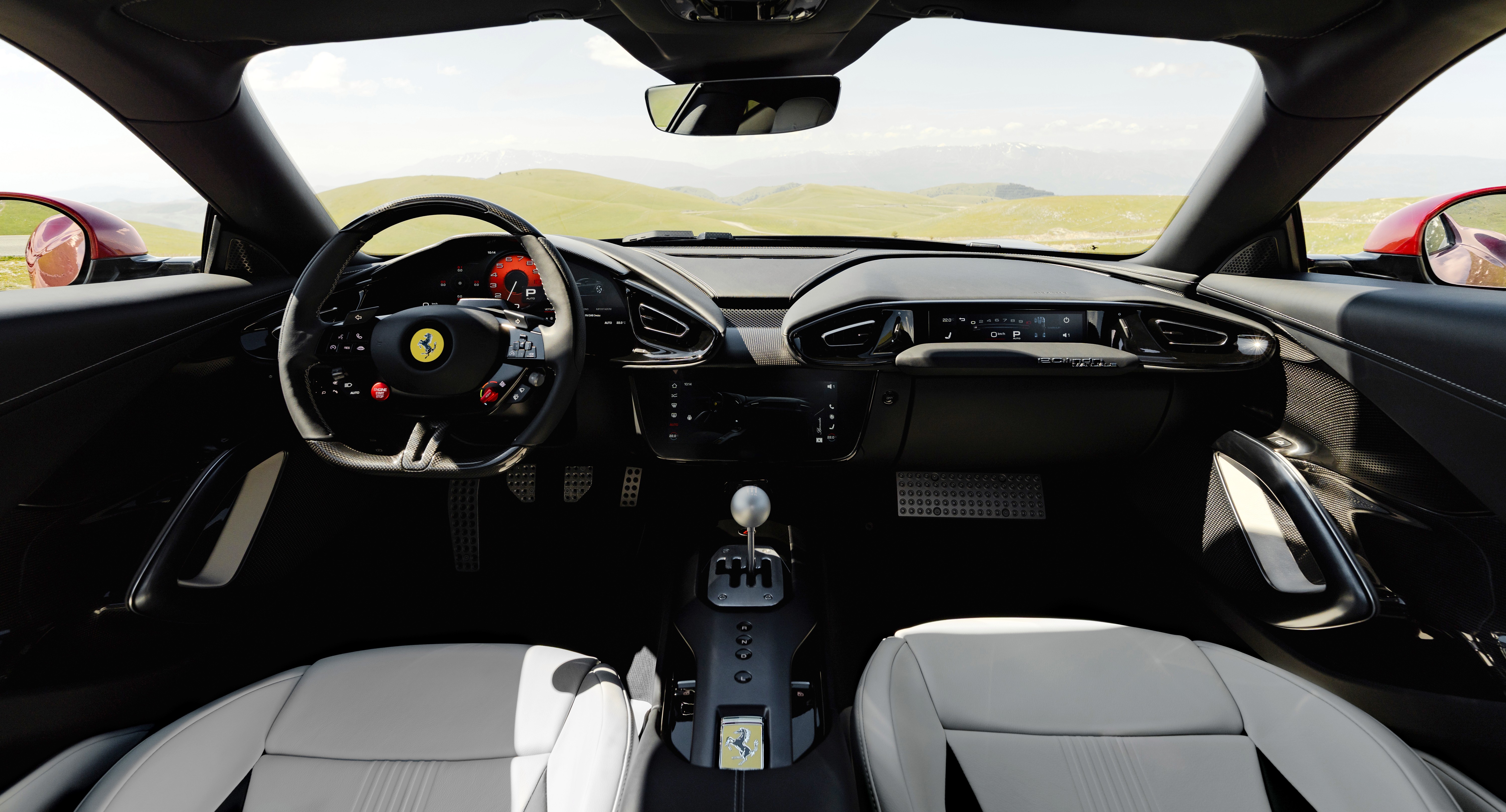 Interior de um carro esportivo Ferrari, com bancos de couro branco, volante preto com o emblema da Ferrari e detalhes vermelhos. O painel é escuro, com um câmbio manual prateado no console central. Ao fundo, uma paisagem montanhosa verde sob céu claro, visível através do para-brisa e dos retrovisores laterais vermelhos