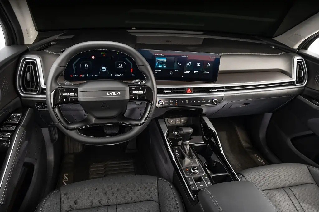 Interior moderno de um carro, com volante multifuncional preto e logo Kia. Painel digital exibe informações do veículo e uma tela multimídia sensível ao toque mostra ícones de aplicativos. O console central possui alavanca de câmbio e botões de controle, bancos de couro escuro e detalhes prateados no painel. A iluminação é suave, destacando o design sofisticado