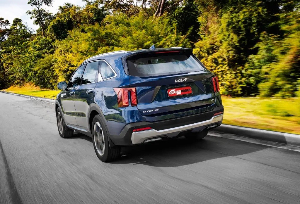 SUV Kia Sorento azul escuro visto por trás, em movimento numa estrada asfaltada, com árvores e vegetação verde ao fundo