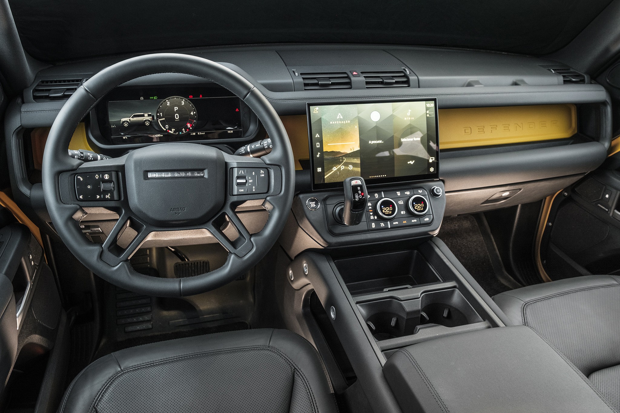 Interior de um carro moderno com volante multifuncional preto, painel digital exibindo um veículo e um velocímetro, e uma tela multimídia central com paisagem. O painel tem detalhes em amarelo e o console central possui porta-copos e apoio de braço. Os bancos são de couro preto.