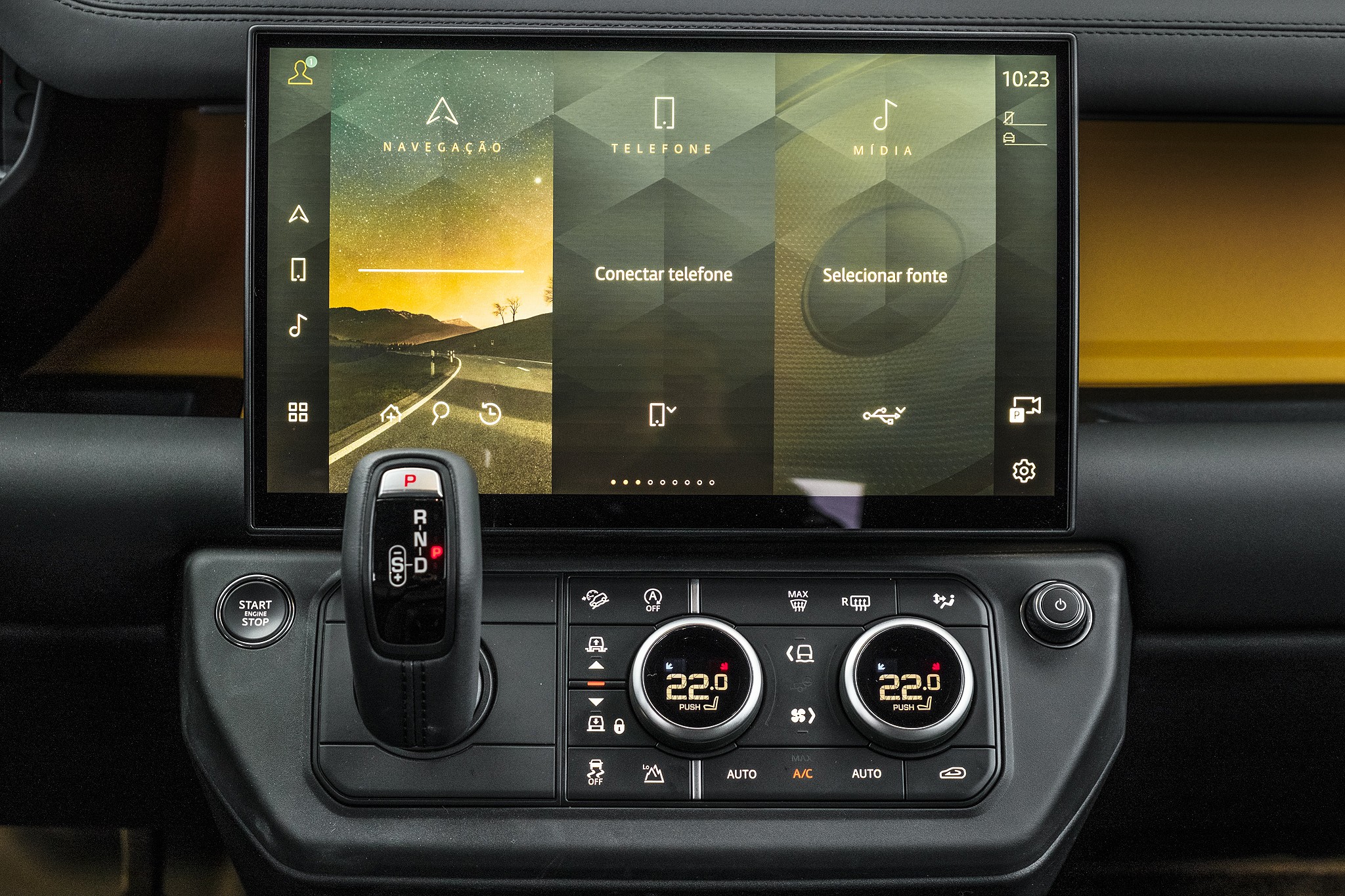 Painel interno de um carro com tela multimídia exibindo opções de navegação, telefone e mídia, com uma estrada ao pôr do sol na tela de navegação. Abaixo, alavanca de câmbio, botão Start Stop e controles de ar condicionado com temperatura de 22 graus