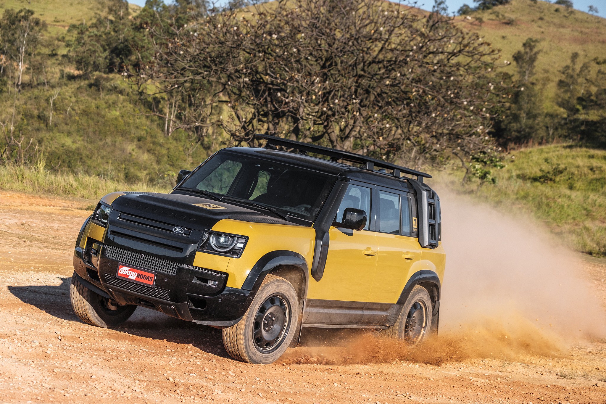 SUV Land Rover Defender amarelo e preto em estrada de terra, levantando poeira. O veículo tem rack de teto e snorkel, com colinas verdes e árvores ao fundo