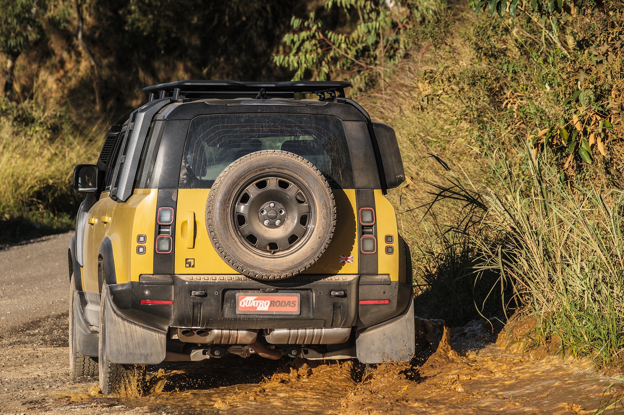 Traseira de um Land Rover Defender amarelo e preto em estrada de terra, com lama espirrando dos pneus. O carro tem estepe na porta traseira, rack de teto e para-barros. Vegetação seca e verde nas laterais da estrada, sob luz solar