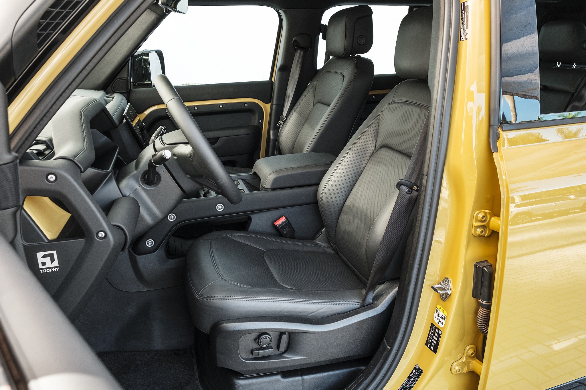 Interior de um carro amarelo com bancos de couro preto, volante e painel escuros. O banco do motorista está em primeiro plano, com cinto de segurança visível. Detalhes dourados no painel e na porta contrastam com o preto, e um emblema Trophy aparece na lateral do console central