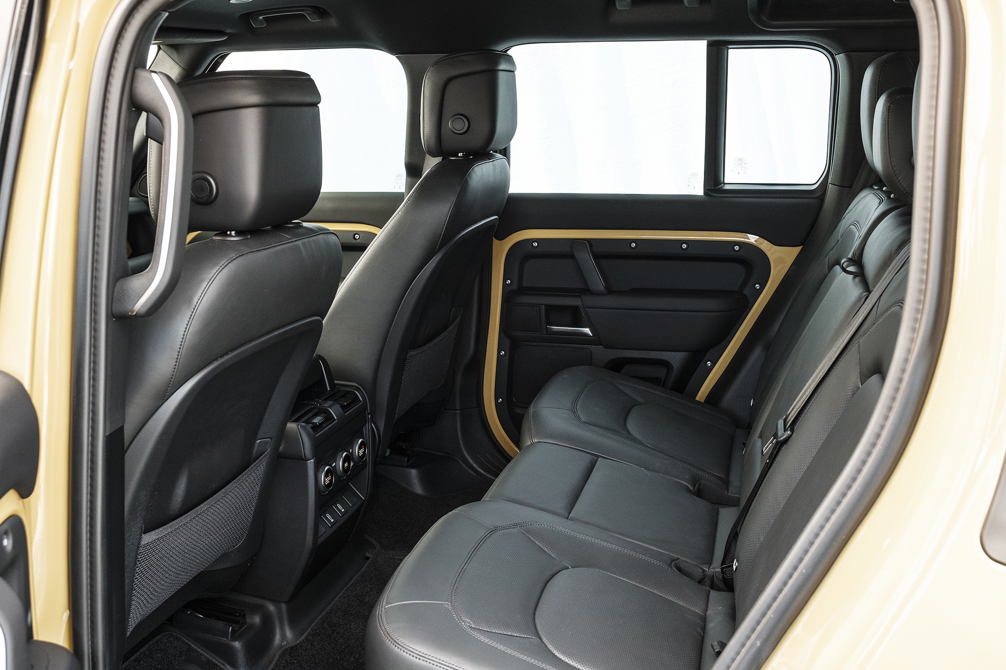 Interior de um carro bege, mostrando os assentos traseiros de couro preto com cintos de segurança e detalhes dourados nas portas