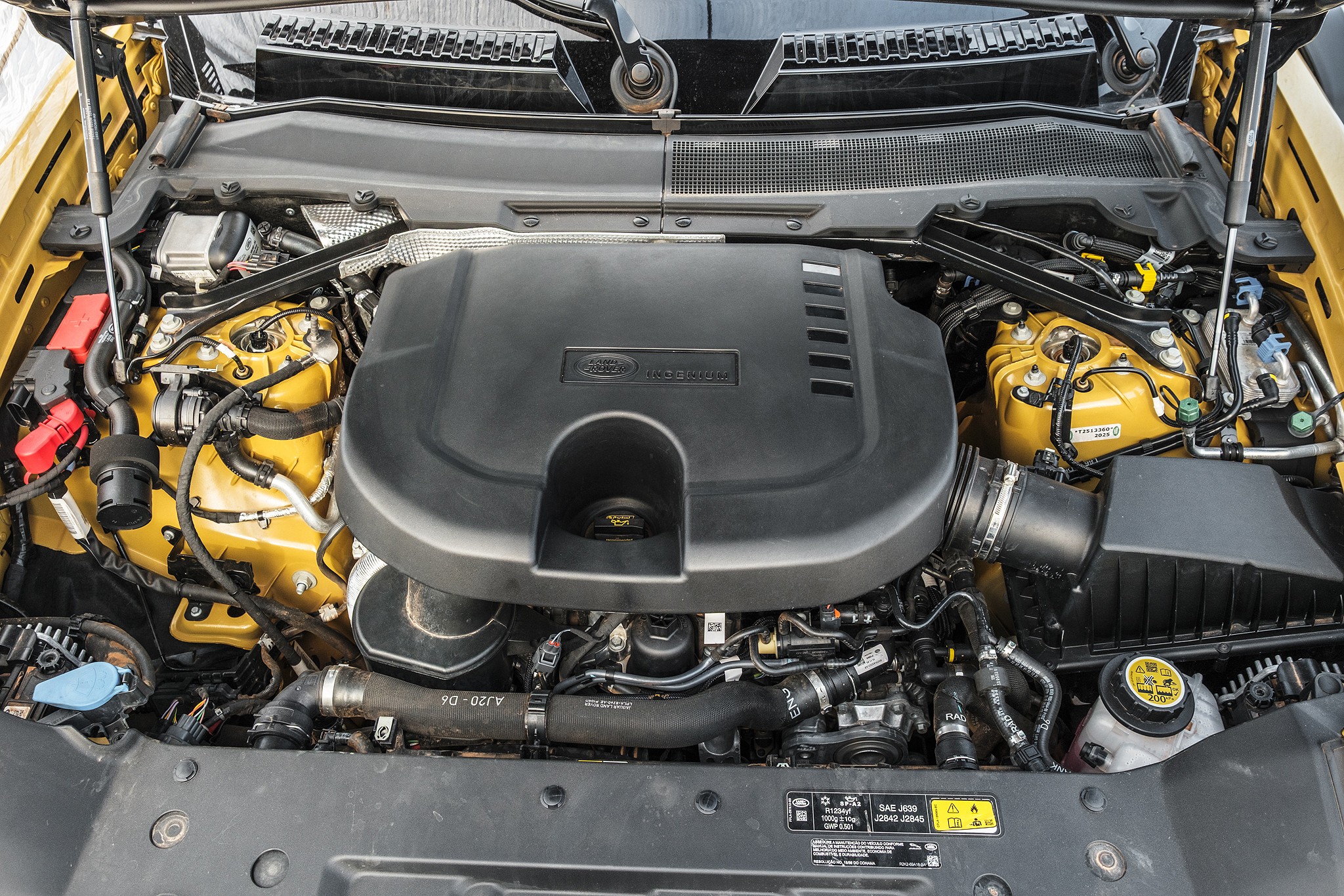 Vista superior do motor de um carro, com a tampa preta do motor em destaque no centro, cercada por componentes amarelos e mangueiras pretas. O capô está aberto, revelando a complexidade do compartimento do motor, com detalhes como o reservatório de fluido de freio e o filtro de ar visíveis. O fundo é um tom de amarelo, indicando a cor do veículo