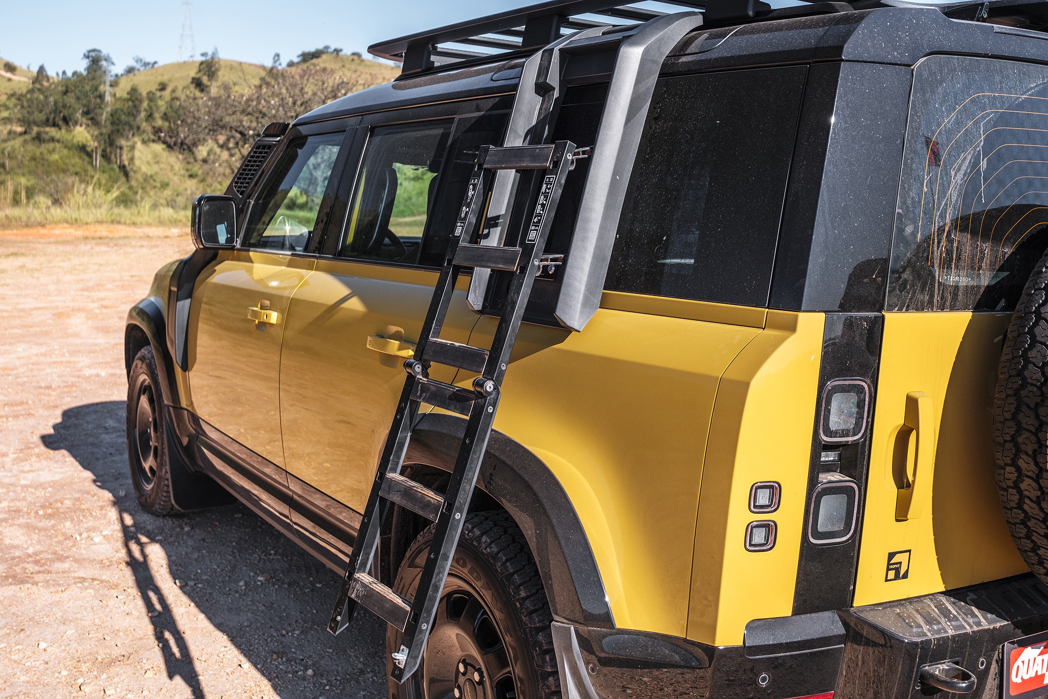 Lateral e traseira de um SUV amarelo com teto preto, rack de teto e escada preta dobrável na lateral. O veículo está estacionado em uma estrada de terra, com colinas e vegetação ao fundo sob um céu azul