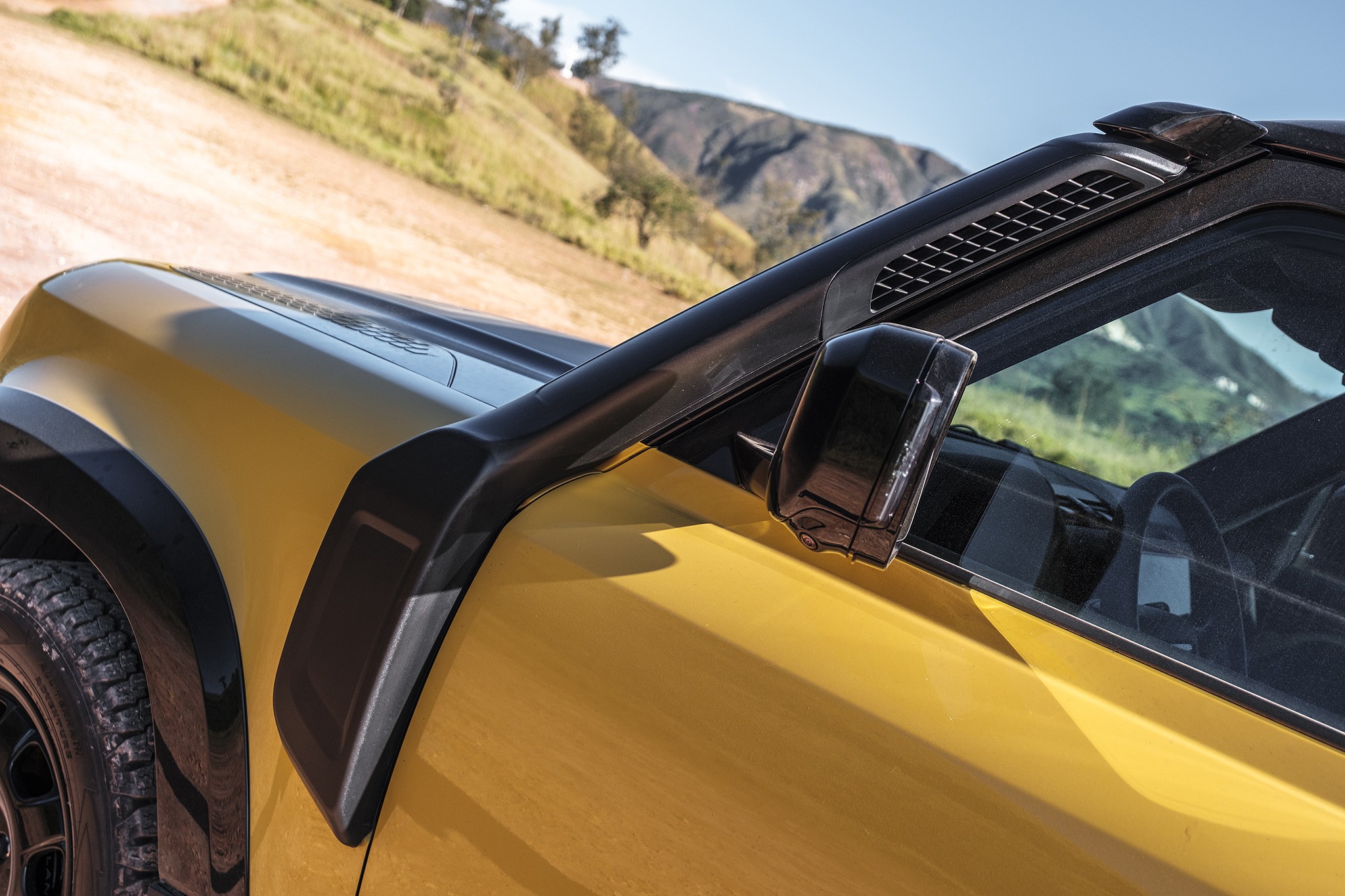 Lateral de um carro amarelo com detalhes pretos, incluindo o para-lama, retrovisor e um snorkel. Ao fundo, uma estrada de terra e montanhas com vegetação rasteira sob céu azul