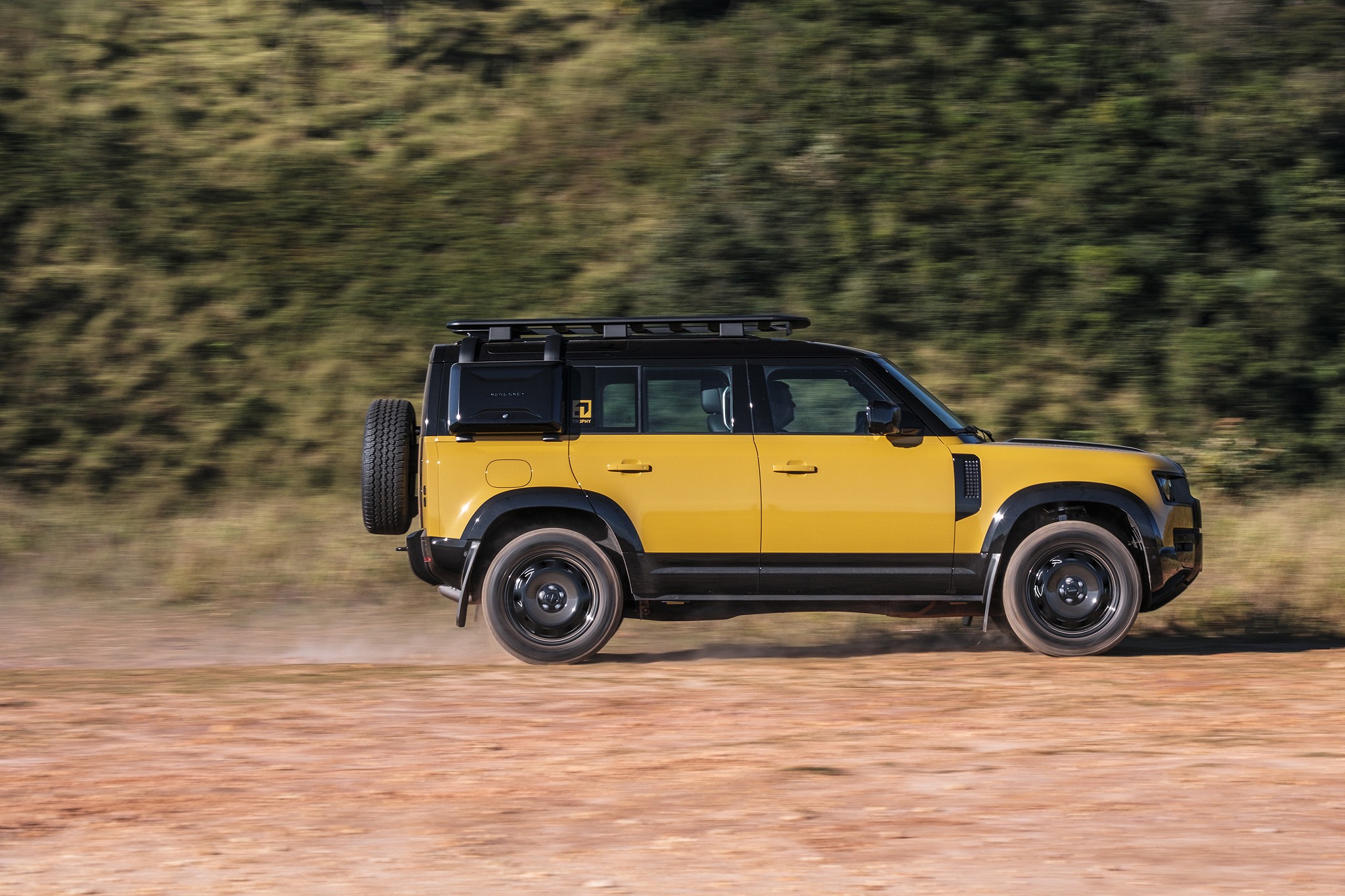 Um Land Rover Defender amarelo e preto em movimento, visto de lado, levantando poeira em uma estrada de terra com vegetação borrada ao fundo