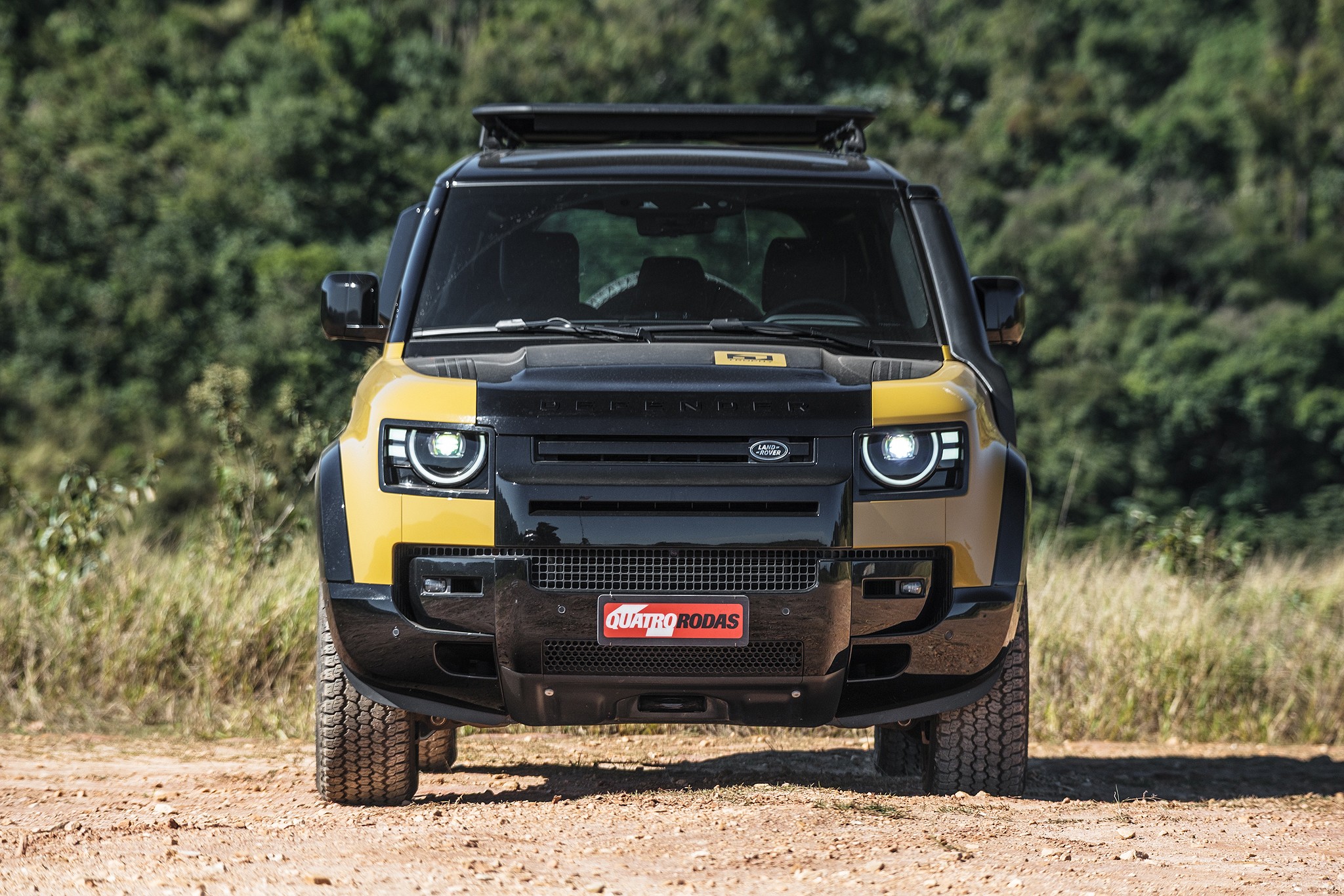 Land Rover Defender amarelo e preto, com bagageiro de teto, parado em estrada de terra. Faróis acesos, grade frontal preta com o logo da Land Rover e placa Quatro Rodas vermelha. Ao fundo, vegetação verde e seca