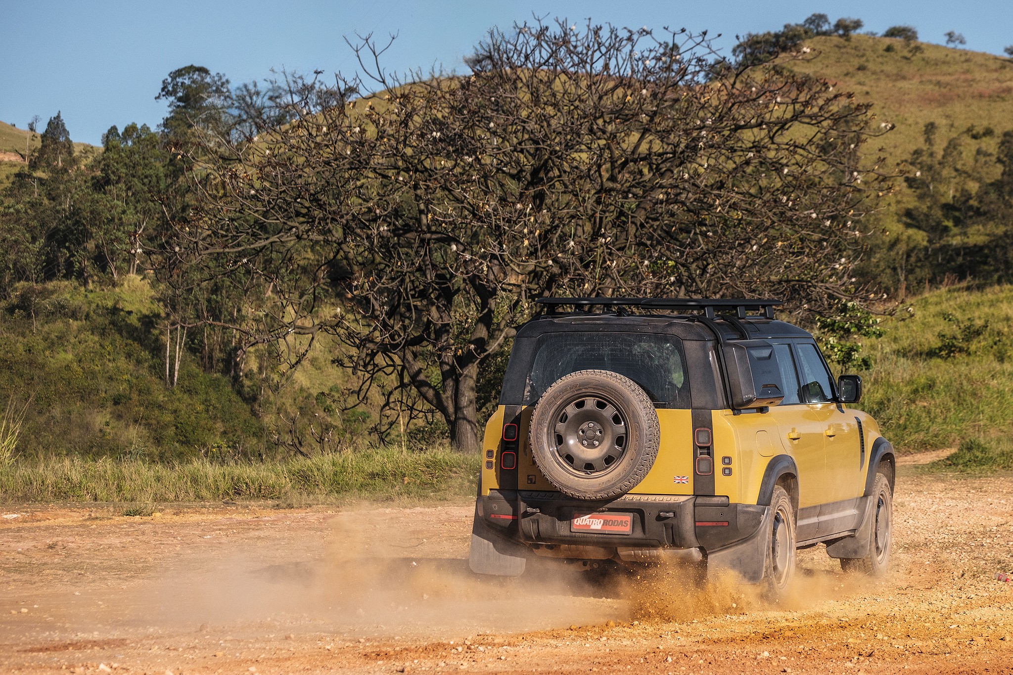 SUV amarelo com teto preto e estepe na traseira, levantando poeira em estrada de terra. Ao fundo, &aacute;rvores secas e colinas verdes sob c&eacute;u azul