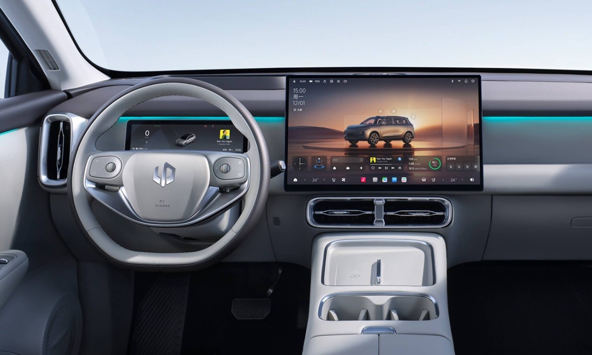 Interior moderno de um carro, com volante cinza claro e um grande painel digital exibindo um SUV prateado em uma paisagem des&eacute;rtica ao p&ocirc;r do sol. H&aacute; detalhes em LED azul no painel e console central com porta-copos.