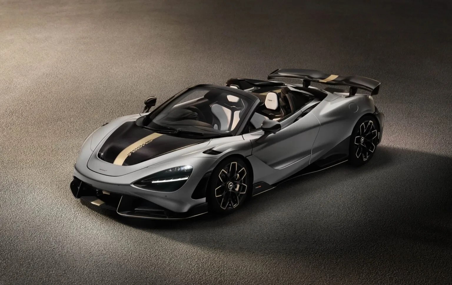 Carro esportivo McLaren 720S Spider cinza metálico com capota abaixada, faixas pretas e douradas no capô e aerofólio, estacionado em piso cinza escuro