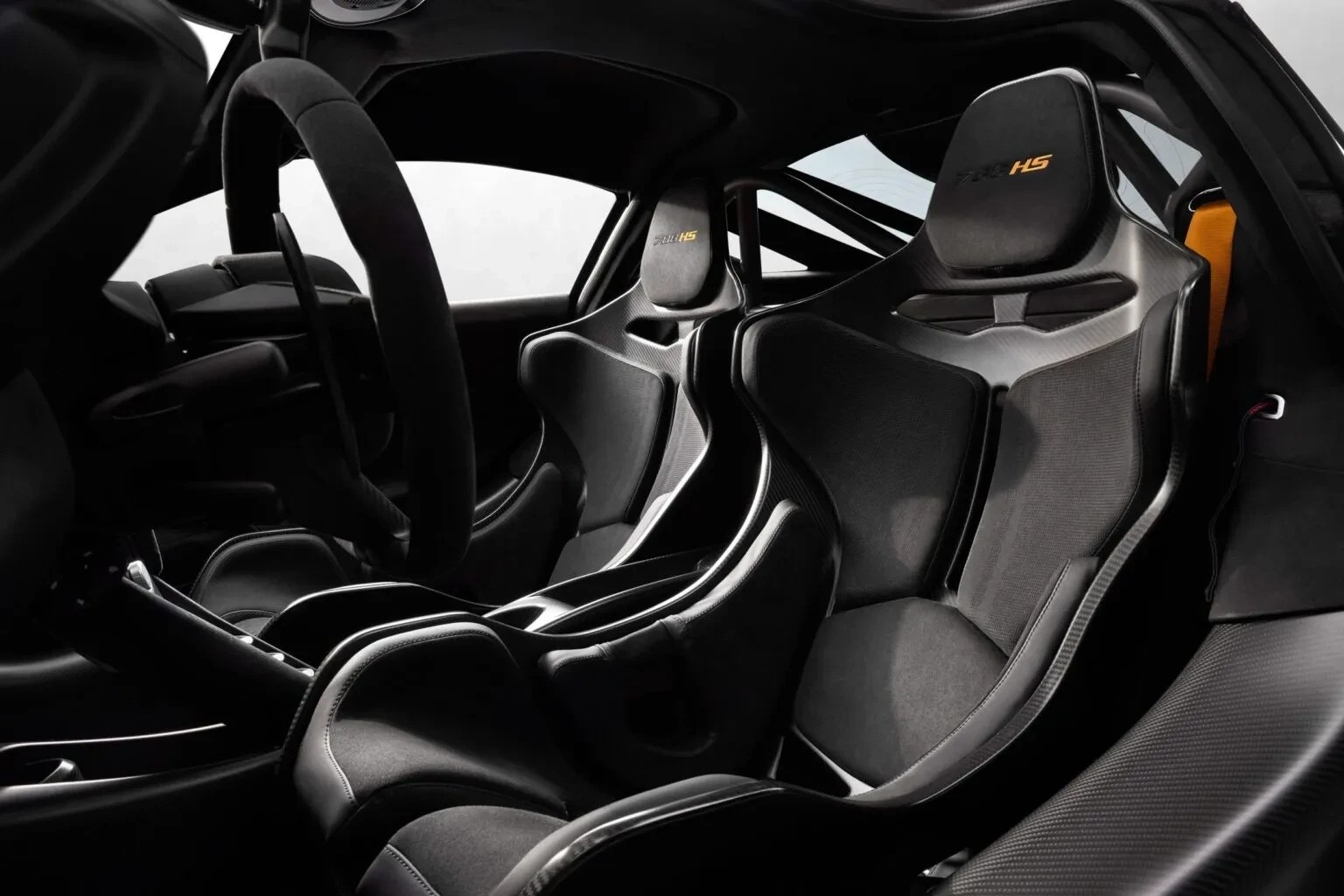 Interior de um carro esportivo com dois bancos concha pretos, volante à esquerda e gaiola de proteção. O encosto de cabeça do banco do passageiro tem o logotipo P1 GTR HS em amarelo. Detalhes em fibra de carbono e um cinto de segurança laranja são visíveis