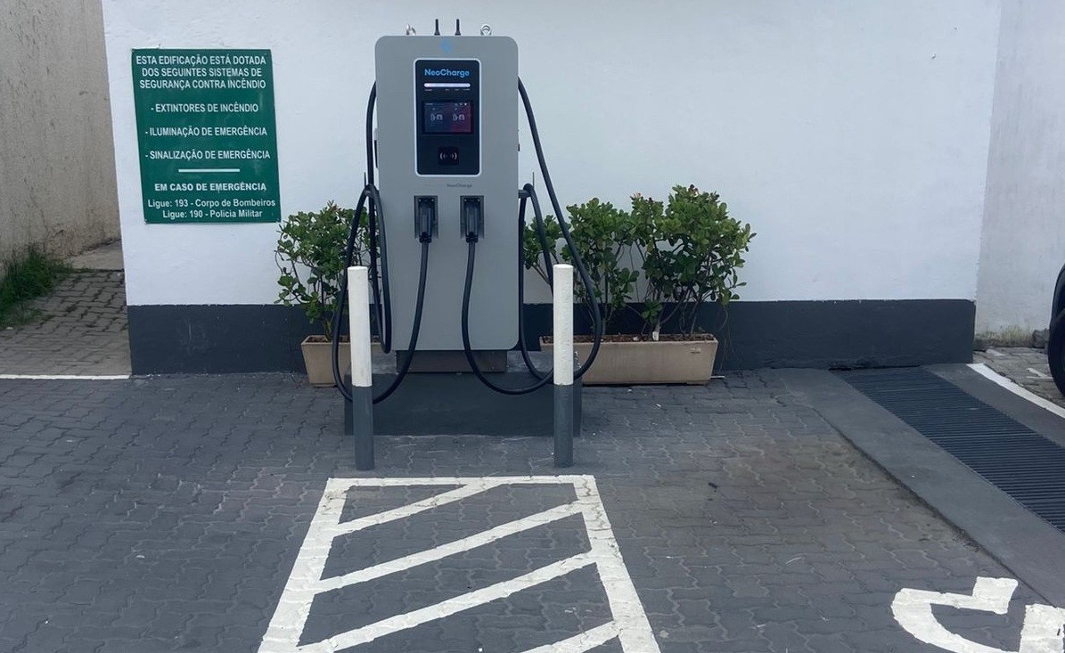 Ponto de recarga para veículos elétricos cinza com duas mangueiras pretas, em uma vaga de estacionamento com sinalização branca no chão. Ao fundo, uma parede branca com uma placa verde de segurança contra incêndio e vasos de plantas.