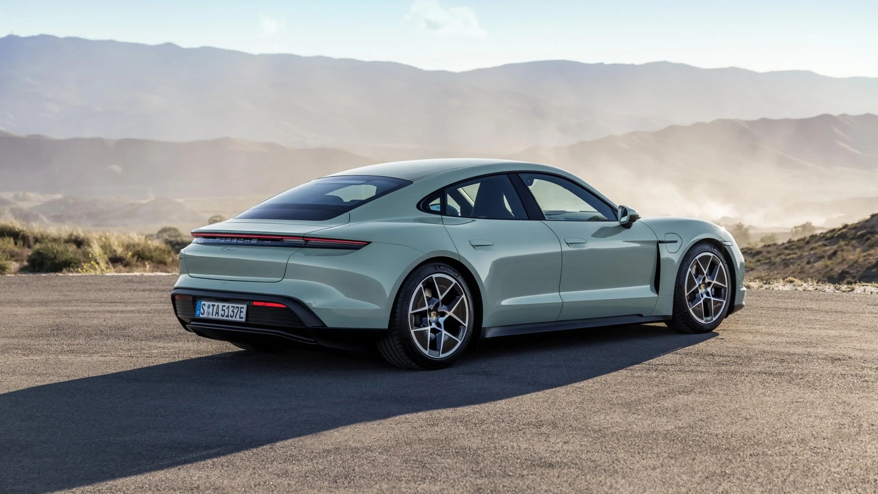 Carro esportivo Porsche Taycan verde-claro estacionado em uma estrada de asfalto, com montanhas e vegetação seca ao fundo sob um céu azul claro