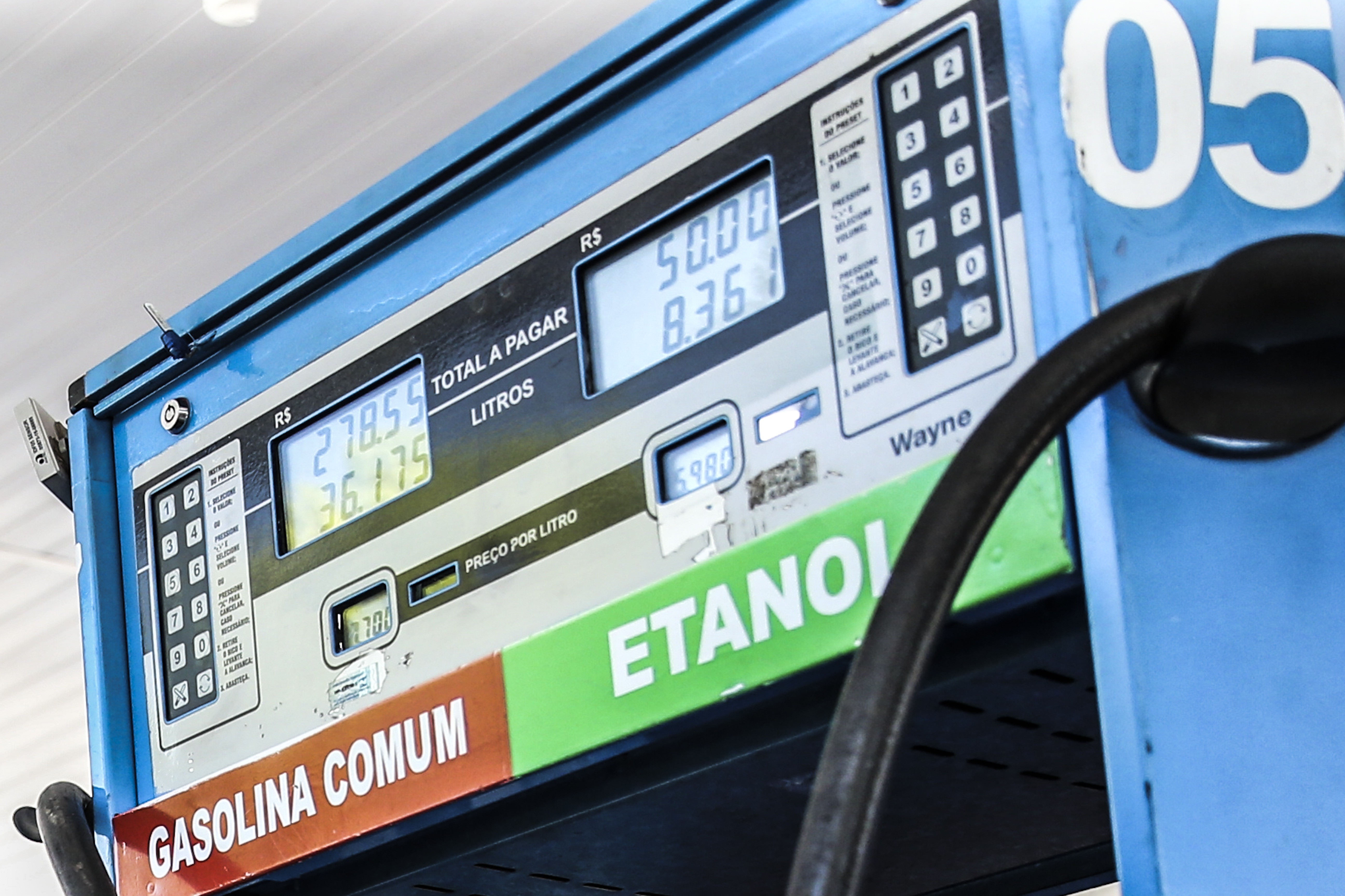 Bomba de combustível azul com visor digital mostrando R$ 278,55 e 36,175 litros de gasolina comum, e R$ 50,00 e 8,36 litros de etanol, com o bico de abastecimento preto à direita