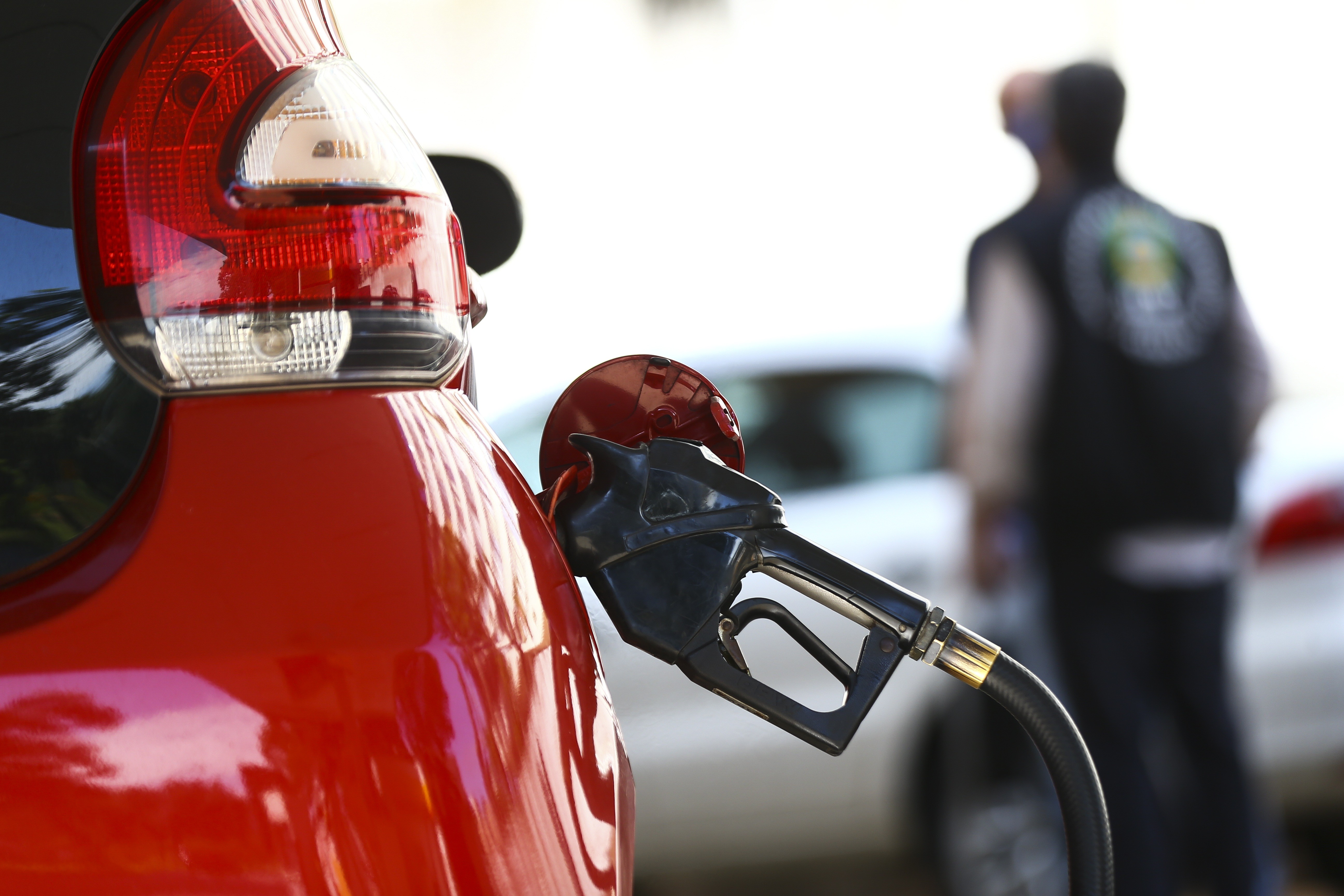 Bico de abastecimento preto inserido no tanque de um carro vermelho, com a traseira do veículo em primeiro plano. Ao fundo, um frentista desfocado de costas e outros carros.