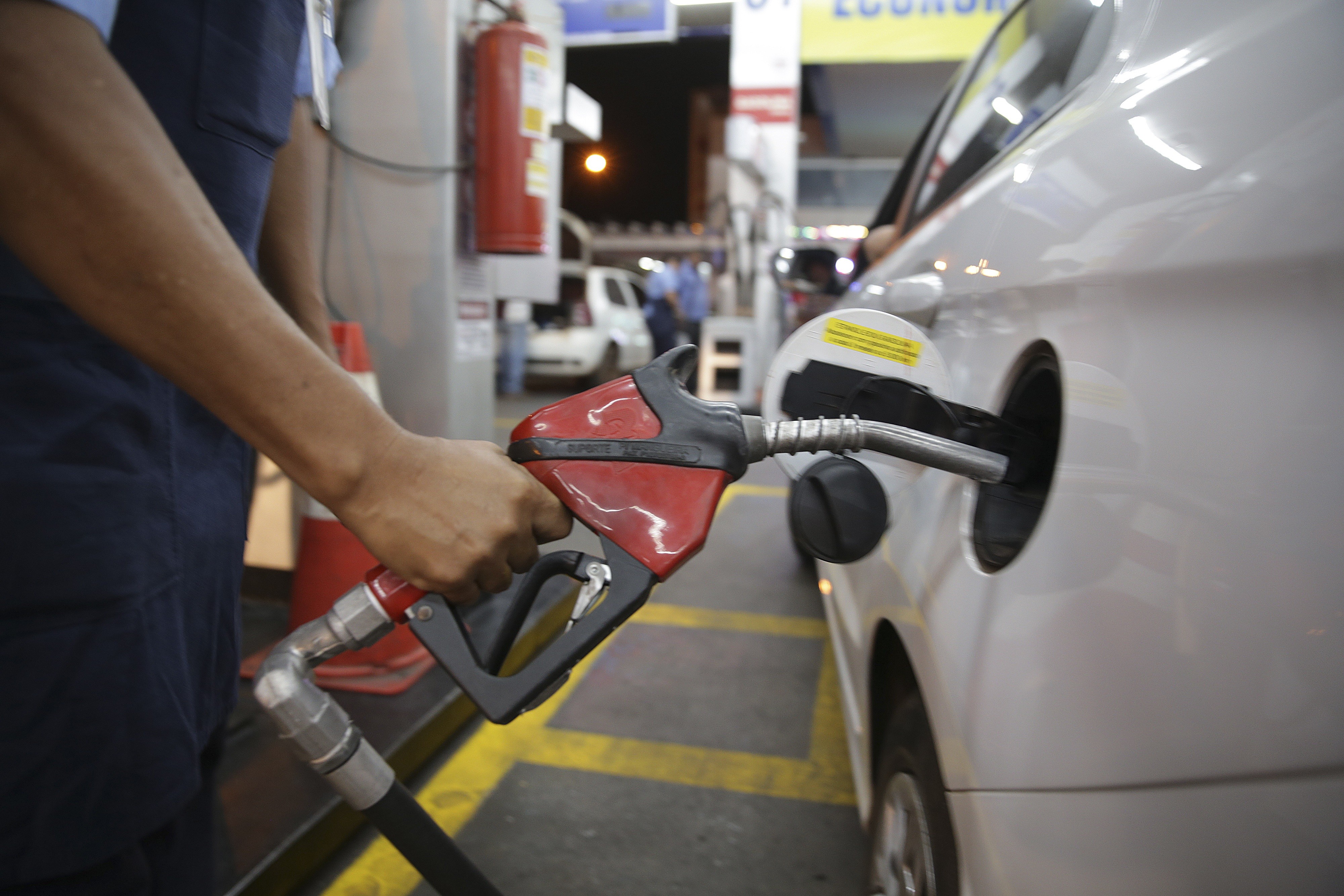 Mão de um frentista segurando um bico de bomba de combustível vermelho e preto, inserido no tanque de um carro branco em um posto de gasolina. Ao fundo, um extintor de incêndio vermelho e luzes noturnas