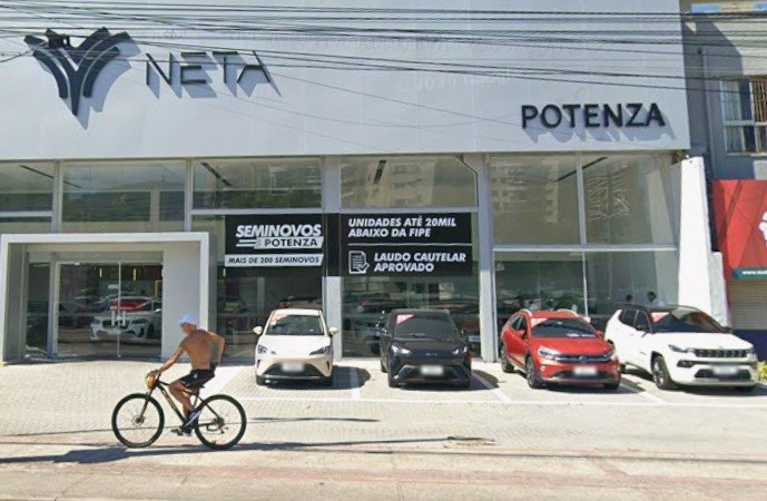 Homem sem camisa e de boné branco pedala uma bicicleta preta em frente à concessionária Neta Potenza, com carros expostos e anúncios de seminovos e laudo cautelar aprovado