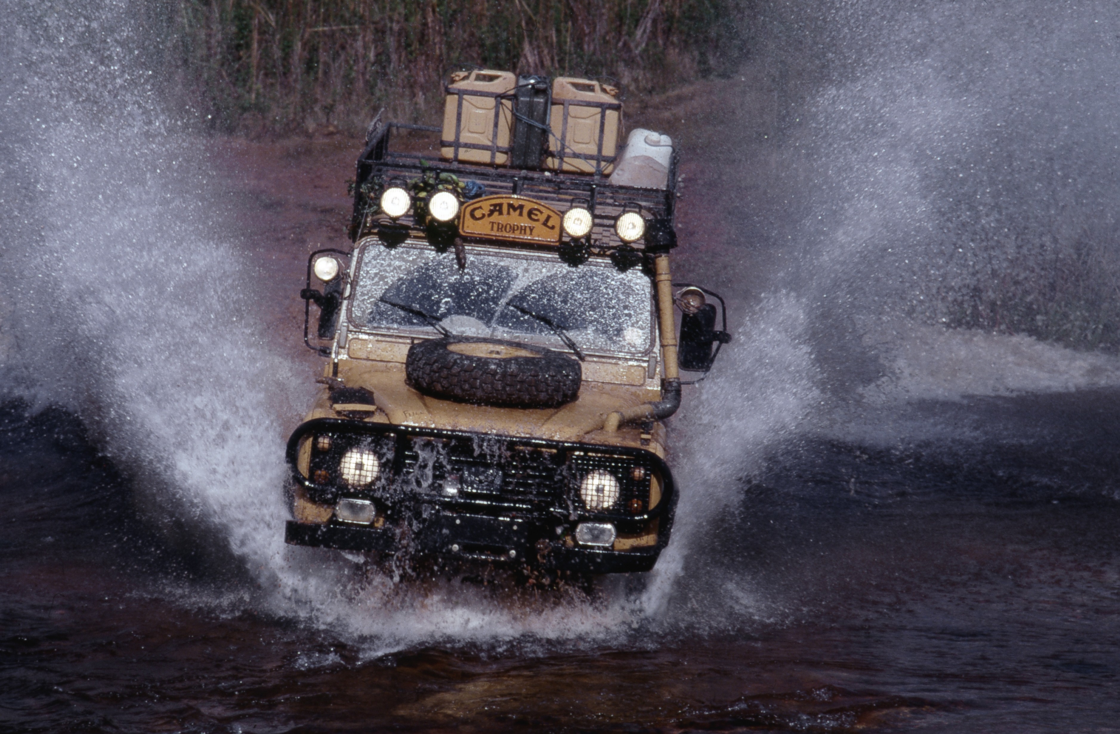 Jipe Land Rover Defender bege com Camel Trophy no teto, atravessando um rio, levantando grandes jatos de água para os lados e sobre o para-brisa