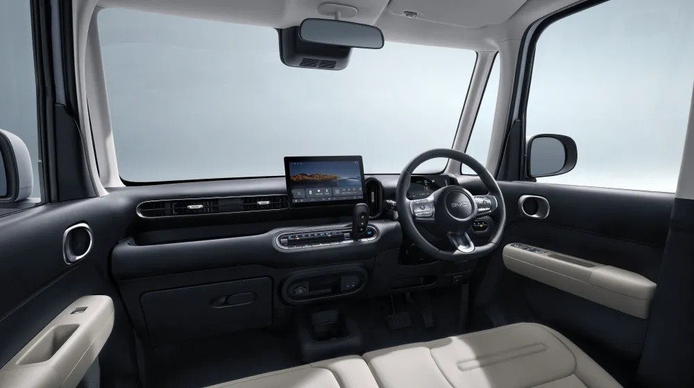 Interior de um carro moderno, com painel escuro e detalhes claros. Volante multifuncional à direita, tela multimídia central exibindo uma paisagem. Bancos de couro bege e espelho retrovisor visível.