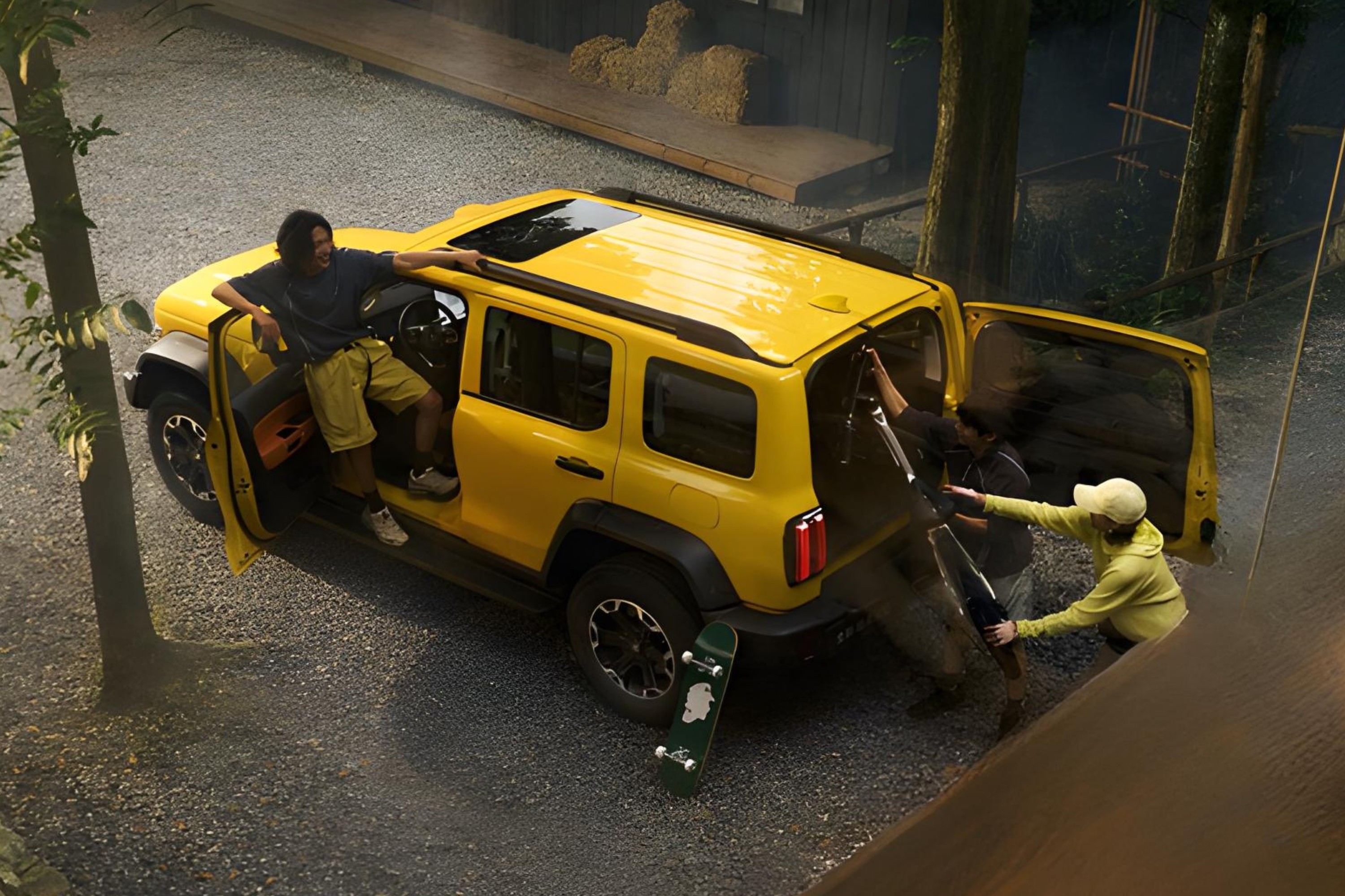 Três pessoas descarregam equipamentos de um SUV amarelo em uma estrada de cascalho. Um homem de camiseta escura e bermuda clara está na porta do motorista, sorrindo. Outras duas pessoas, uma de boné e blusa amarela, tiram itens do porta-malas. Um skate verde está no chão, perto da roda traseira. Ao fundo, árvores e uma construção de madeira com fardos de feno