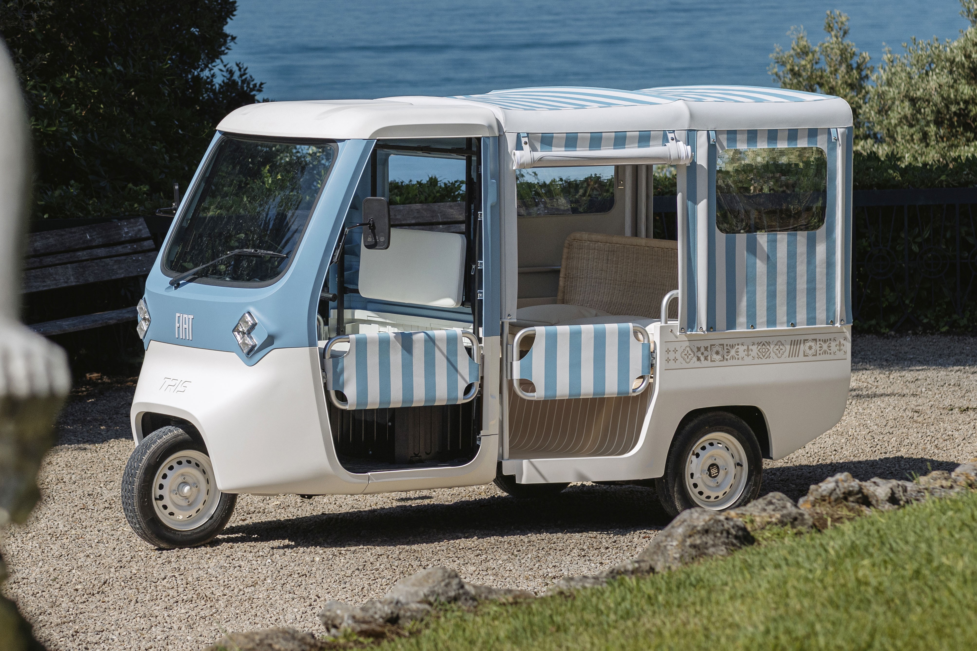 Um veículo elétrico de três rodas, branco e azul-claro, com teto e laterais listrados em azul e branco, estacionado em um caminho de cascalho. O mar azul e a vegetação verde aparecem ao fundo