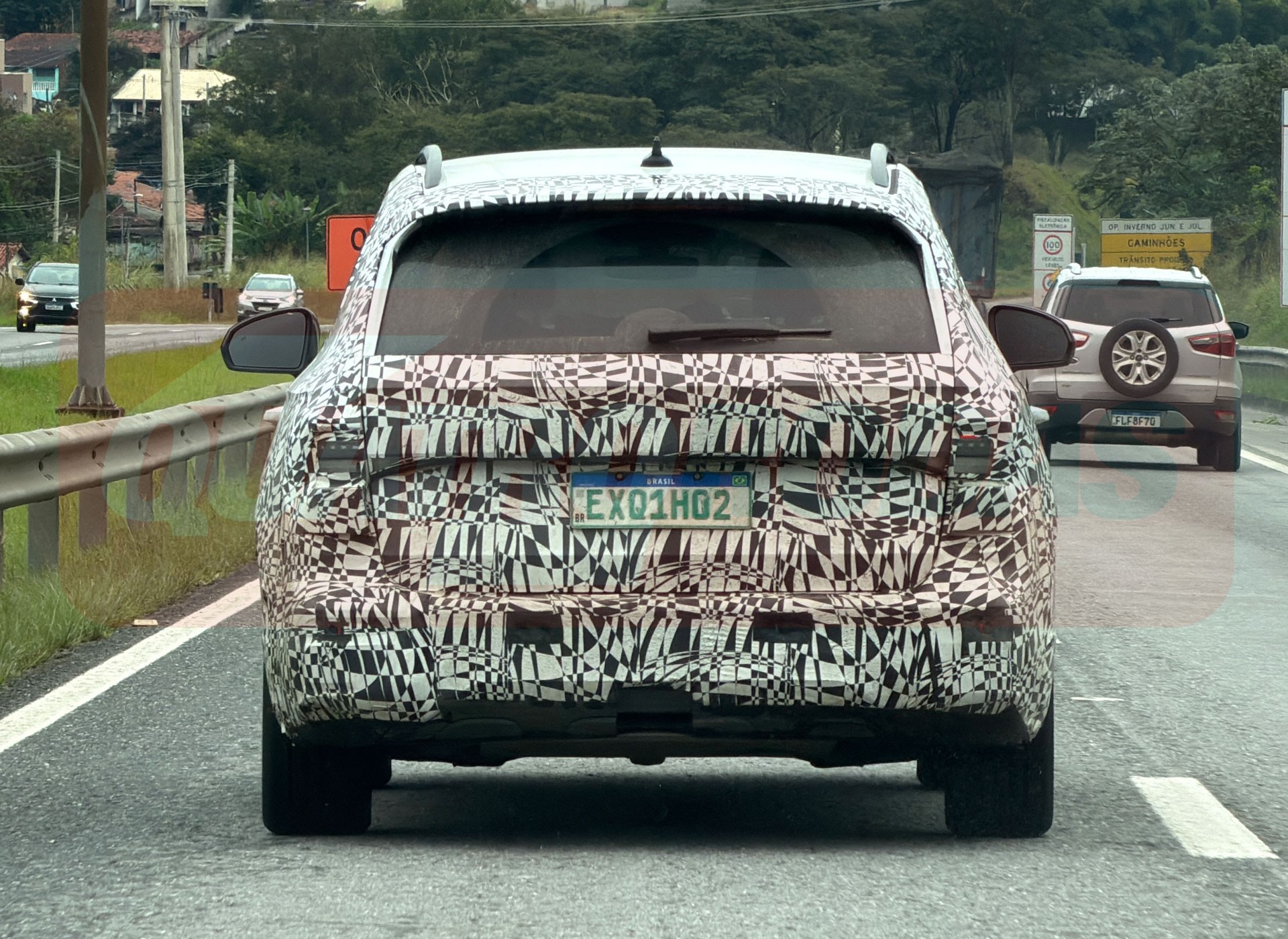 Traseira de um carro camuflado com adesivos pretos e brancos, placa EXQ1H02, em uma estrada. À direita, um SUV prata com estepe na tampa traseira.