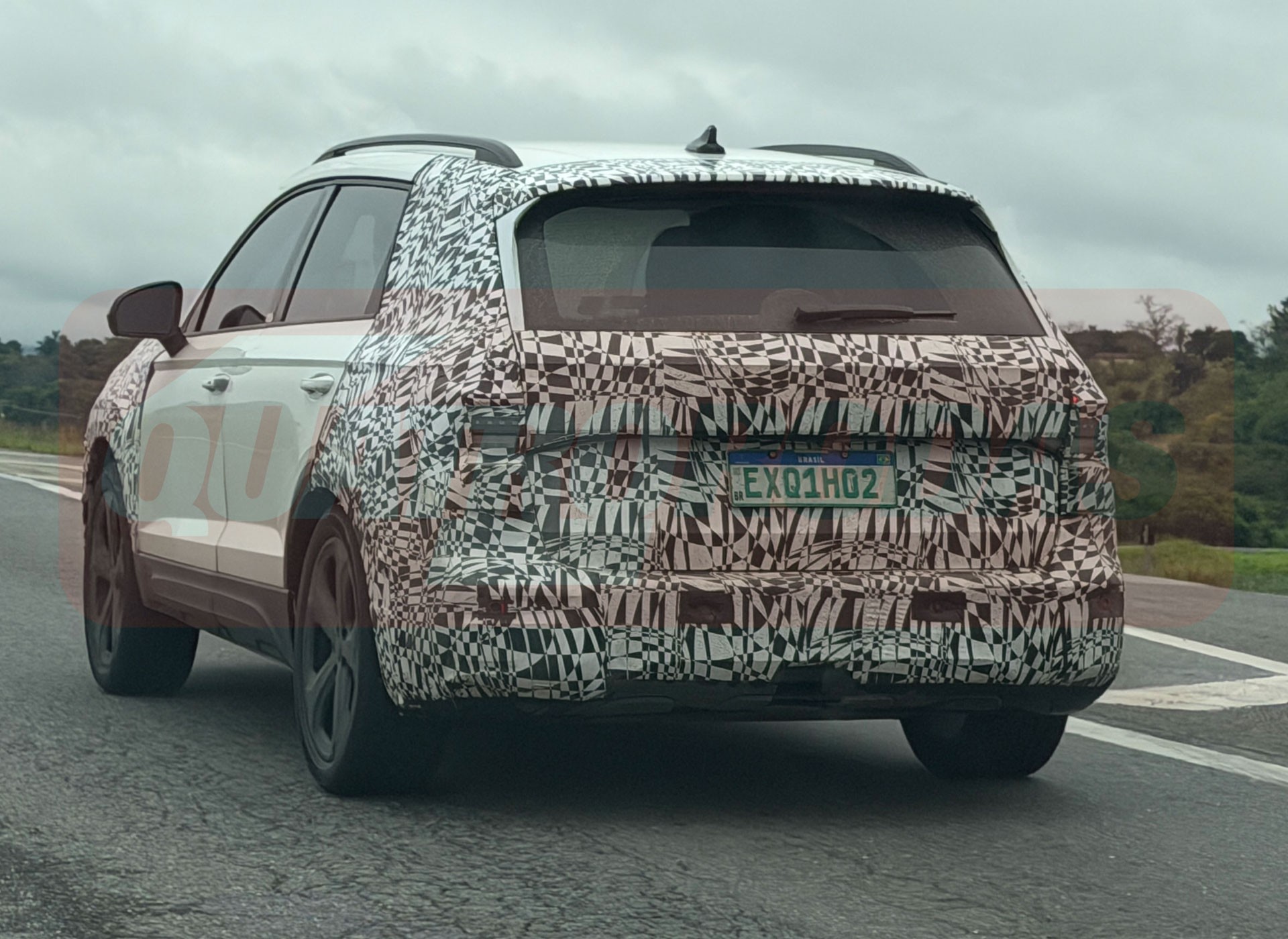 SUV branco camuflado com adesivos pretos e brancos, visto de traseira e lateral, trafegando em estrada sob céu nublado. A placa de teste é verde com letras brancas EXQ1H02