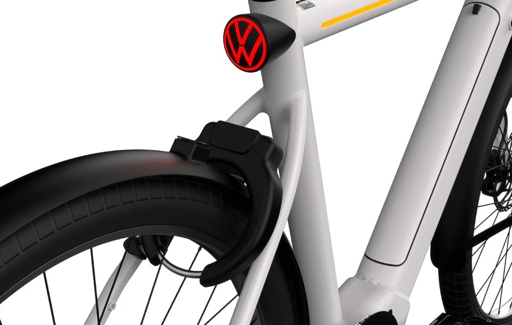 Detalhe da parte traseira de uma bicicleta elétrica branca, com para-lama preto e pneu com banda de rodagem. Acima do para-lama, uma lanterna traseira vermelha com o logotipo da Volkswagen em preto. Um cadeado em U preto está preso ao quadro, próximo à roda